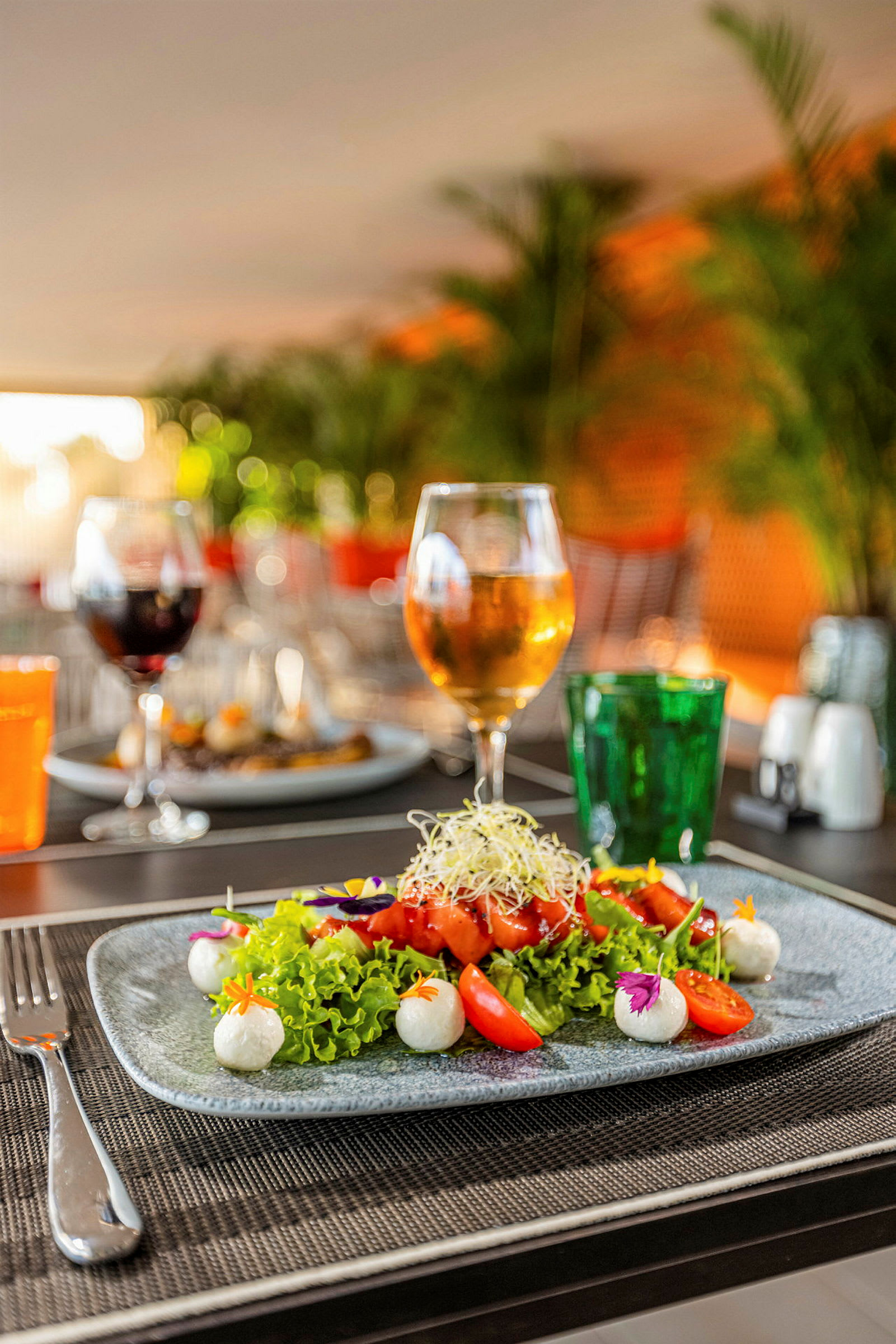 En tallrik med färgglad sallad, tomater och bollar av mozzarella står på ett dukat bord. Bakgrunden visar ett glas med dryck och gröna växter. (AI-genererad text)