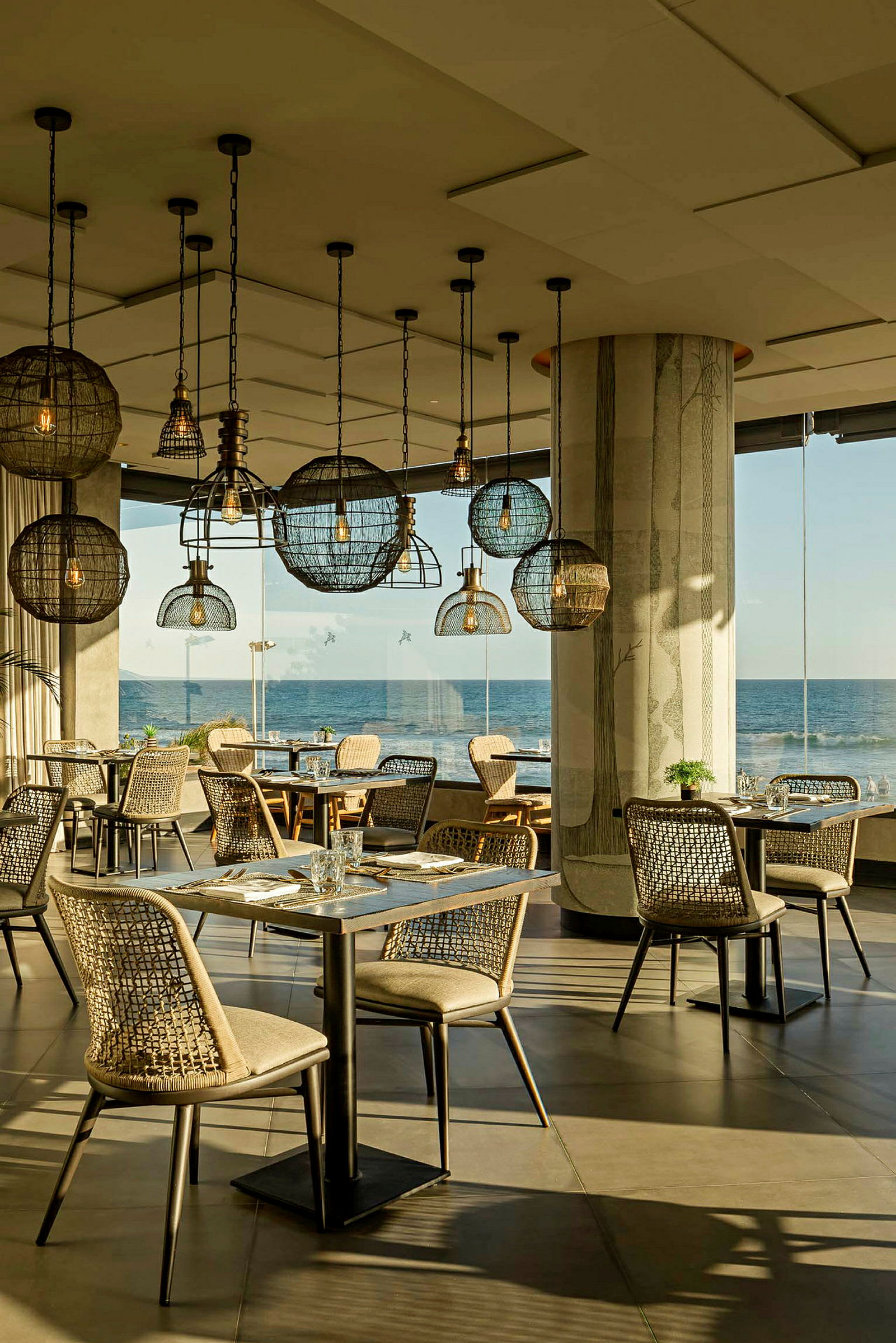 En restaurang med modern inredning, flera matbord och stolar samt dekorativa lampor i taket. Fönster med utsikt över havet. (AI-genererad text)
