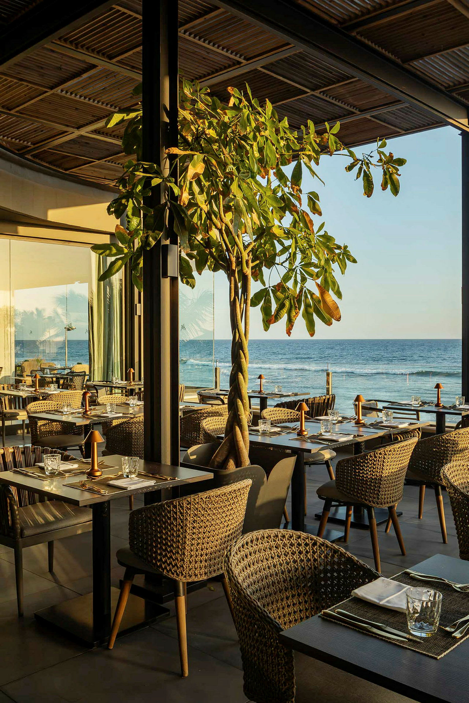 En restaurang med utsikt över havet, där bord och stolar är uppställda i en öppen miljö med naturliga inslag som ett träd. (AI-genererad text)