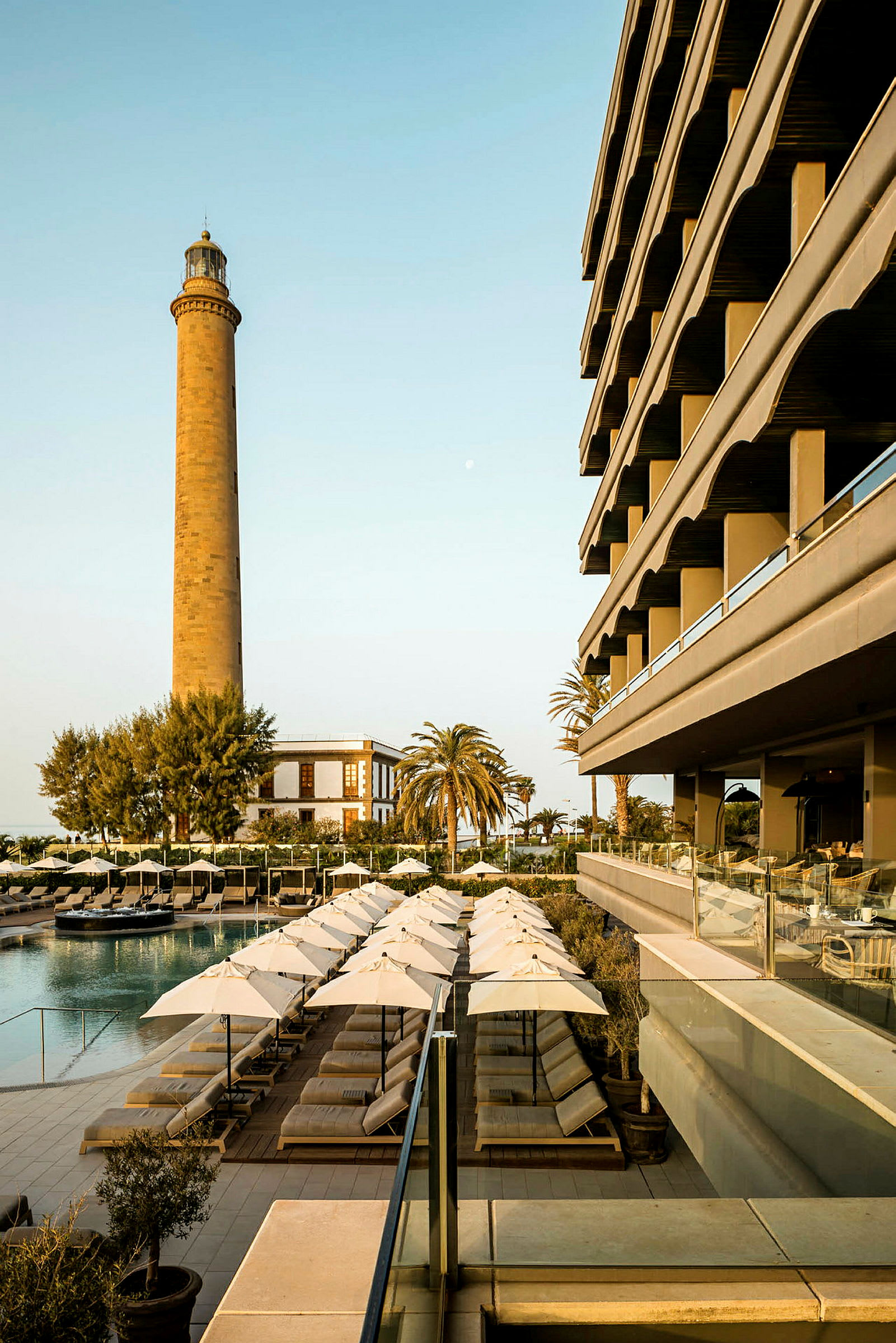 Bild av en hotellbyggnad bredvid en pool med solstolar och parasoller. En fyr och en palm syns i bakgrunden. (AI-genererad text)