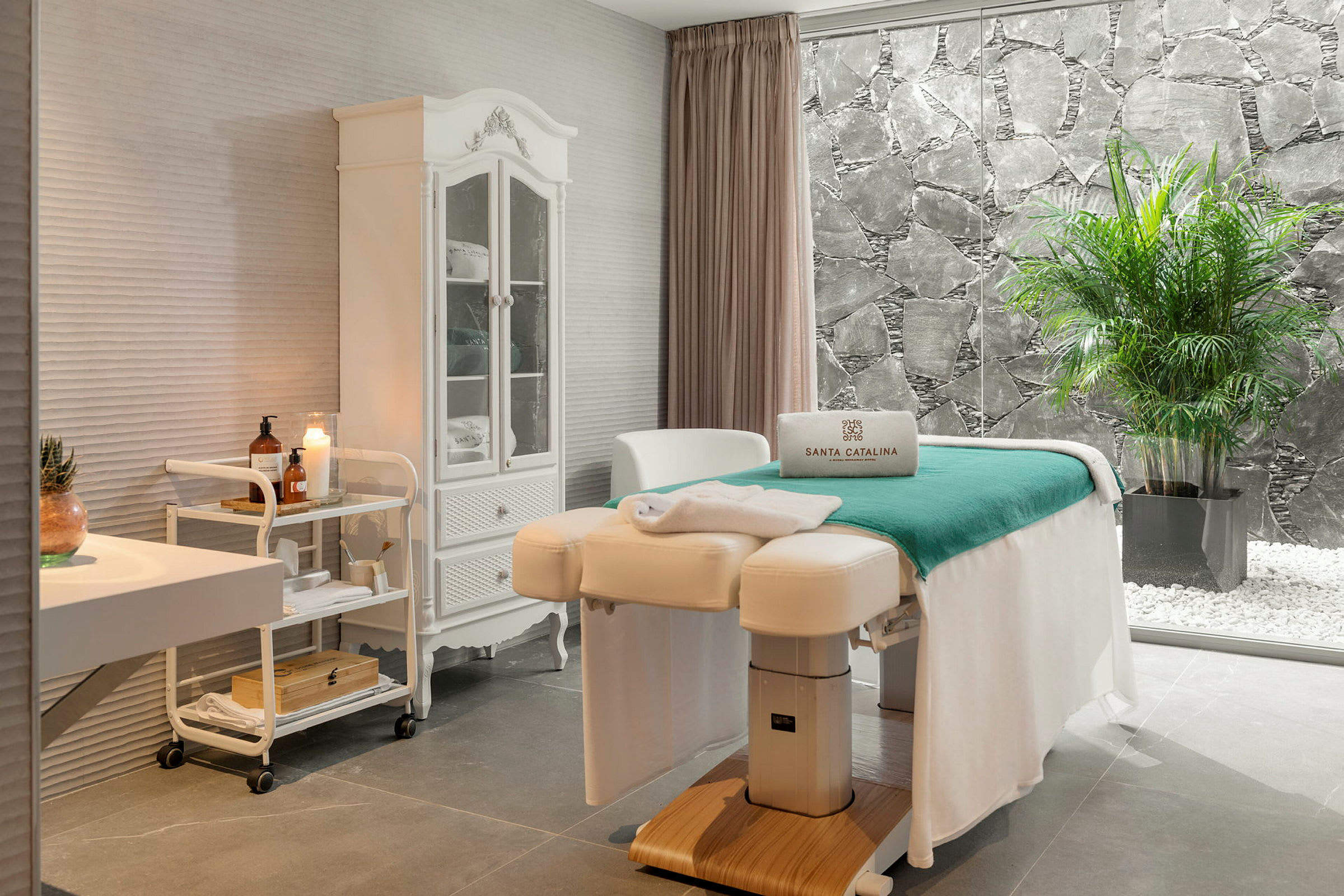 Ett stilrent spa-rum med massagebänk, vit inredning och ett stort fönster med utsikt mot en stenmur och gröna växter. (AI-genererad text)