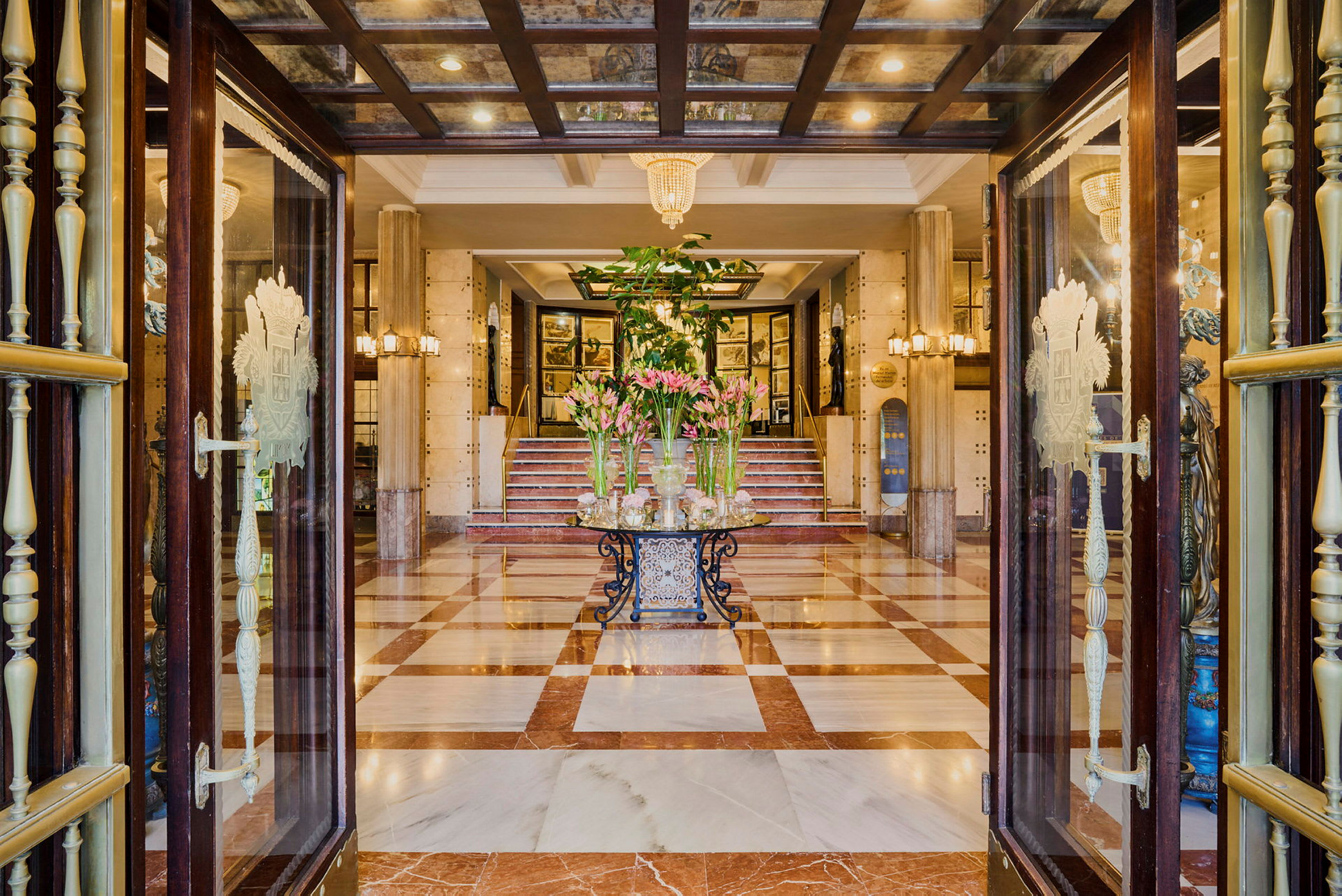 Ett hotellobby med öppna dörrar, marmorgolv och en dekorativ blomsteruppsättning i mitten. (AI-genererad text)