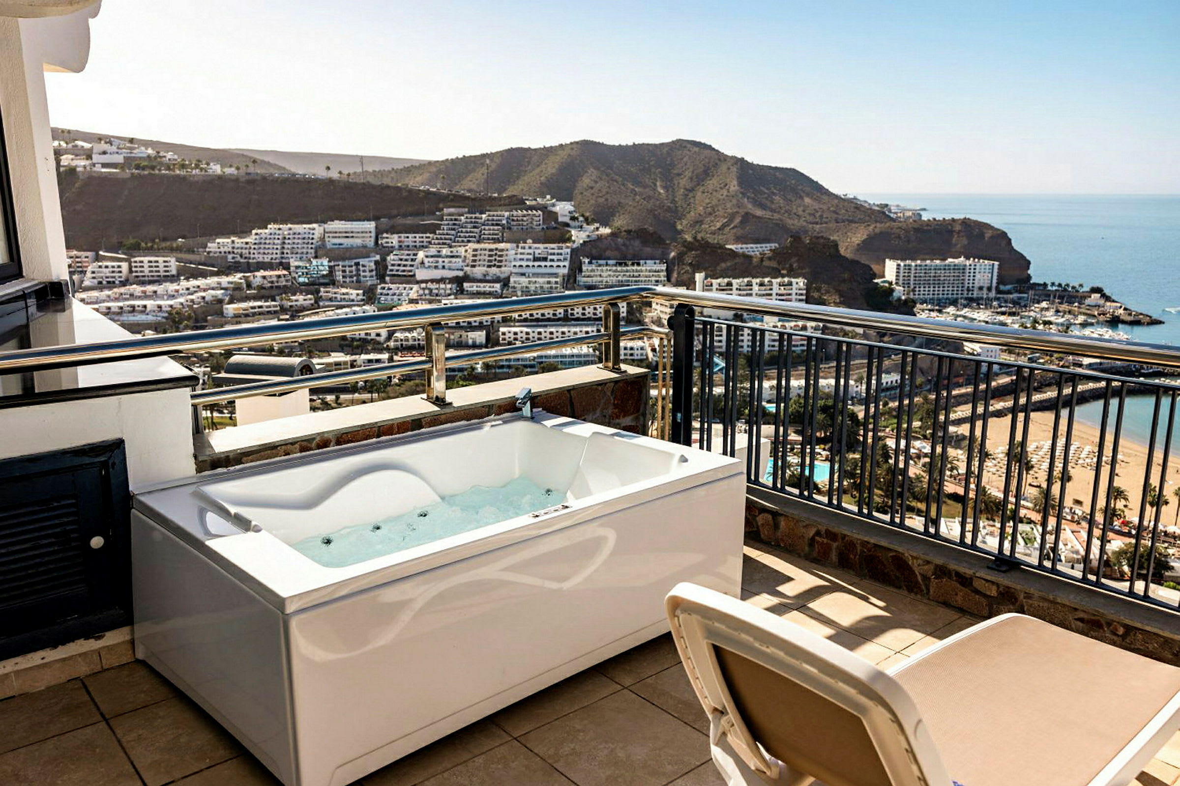 En terrass med bubbelbad och utsikt över en bukt med hotell och strand. (AI-genererad text)