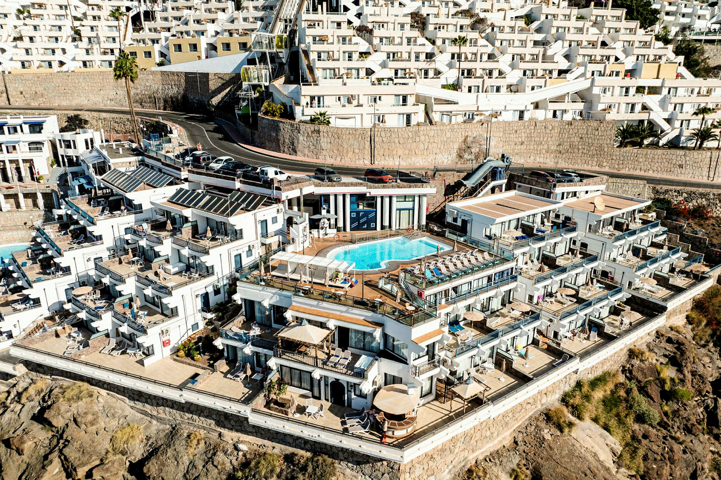 Vy över en flera våningar hög byggnad med en pool och terrasser vid havet. (AI-genererad text)