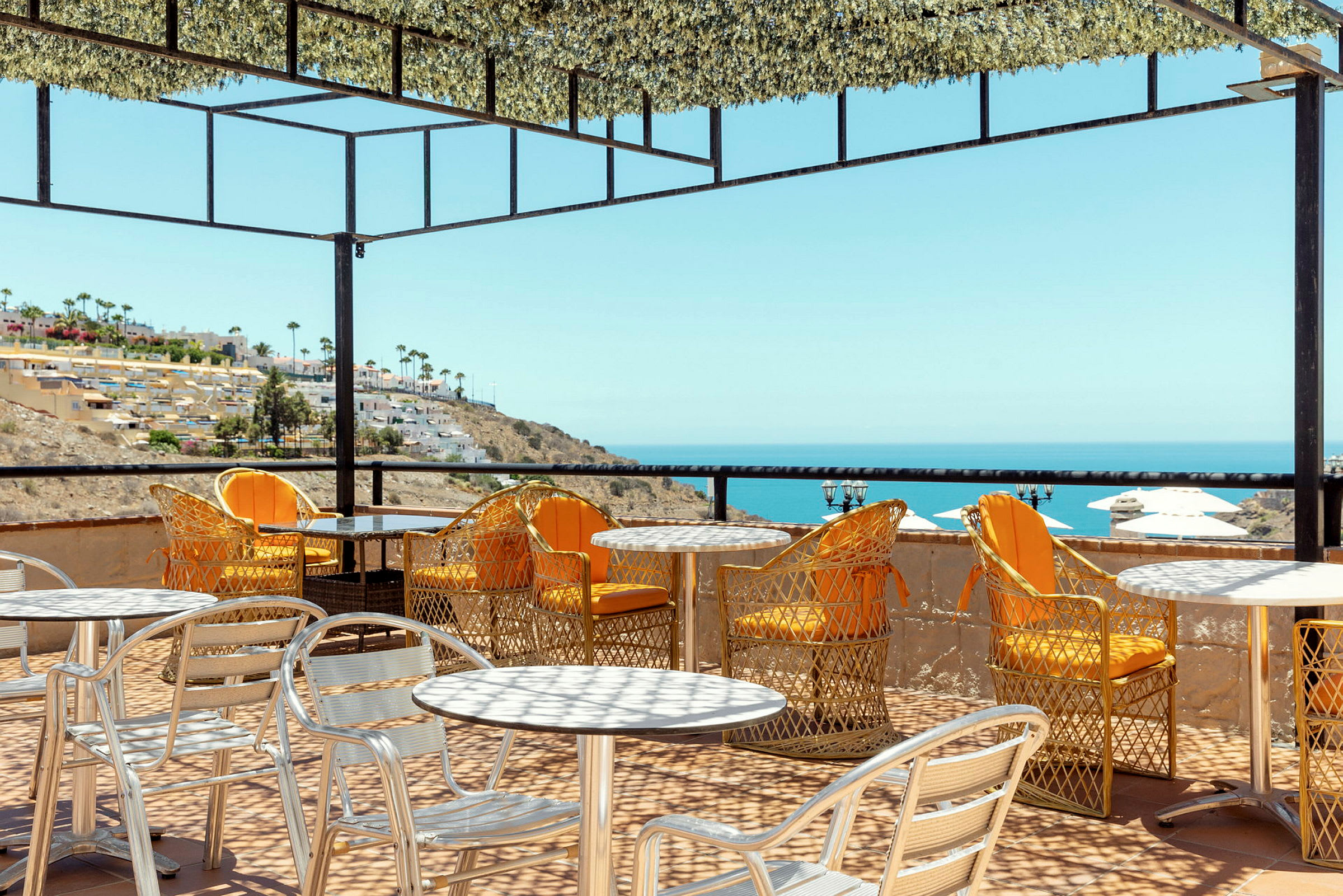 Uteservering med bord och stolar på en terrasse med utsikt över havet och en sluttning med byggnader. (AI-genererad text)