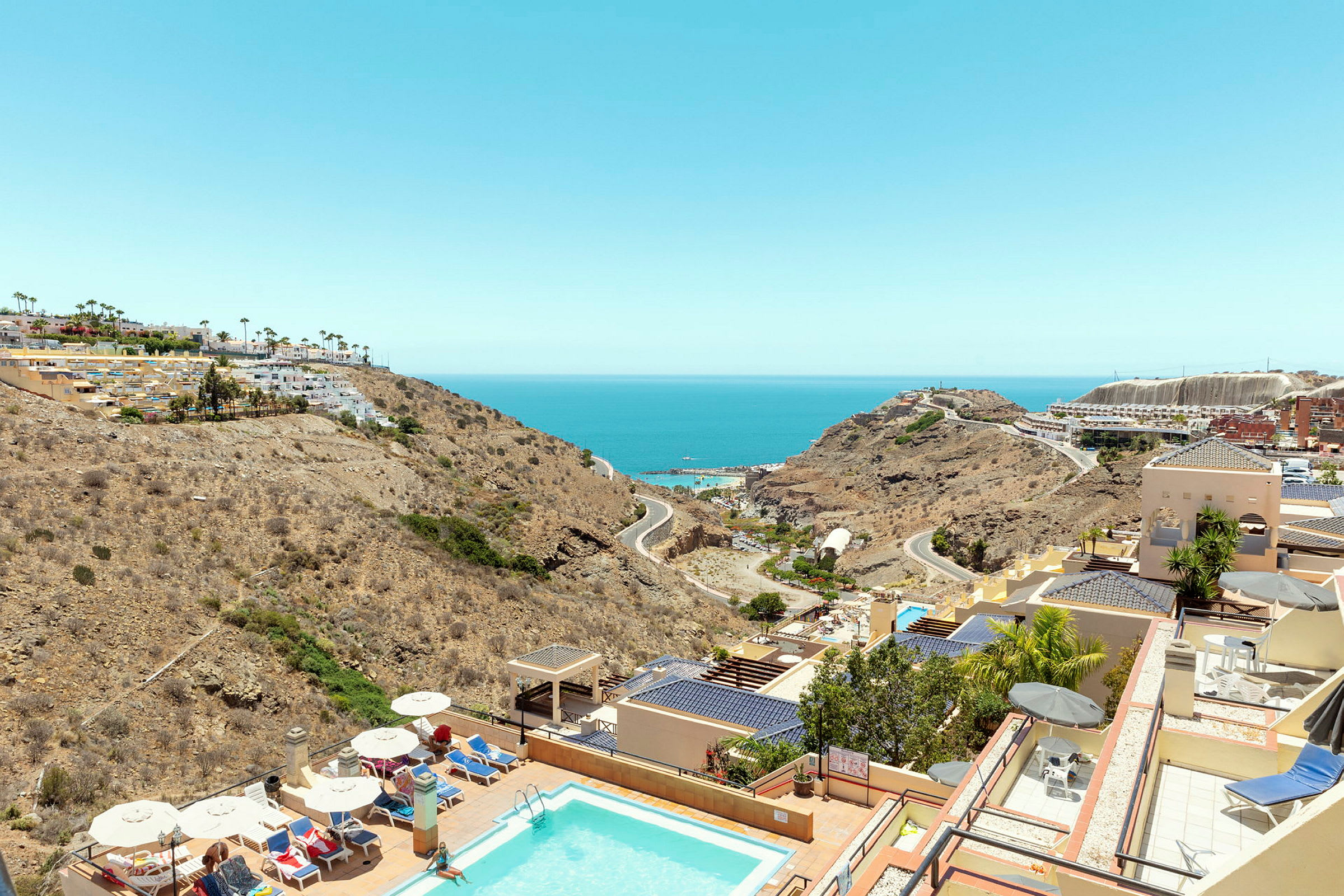 Vy över en pool, hotellbyggnader och landskap mot havet. (AI-genererad text)