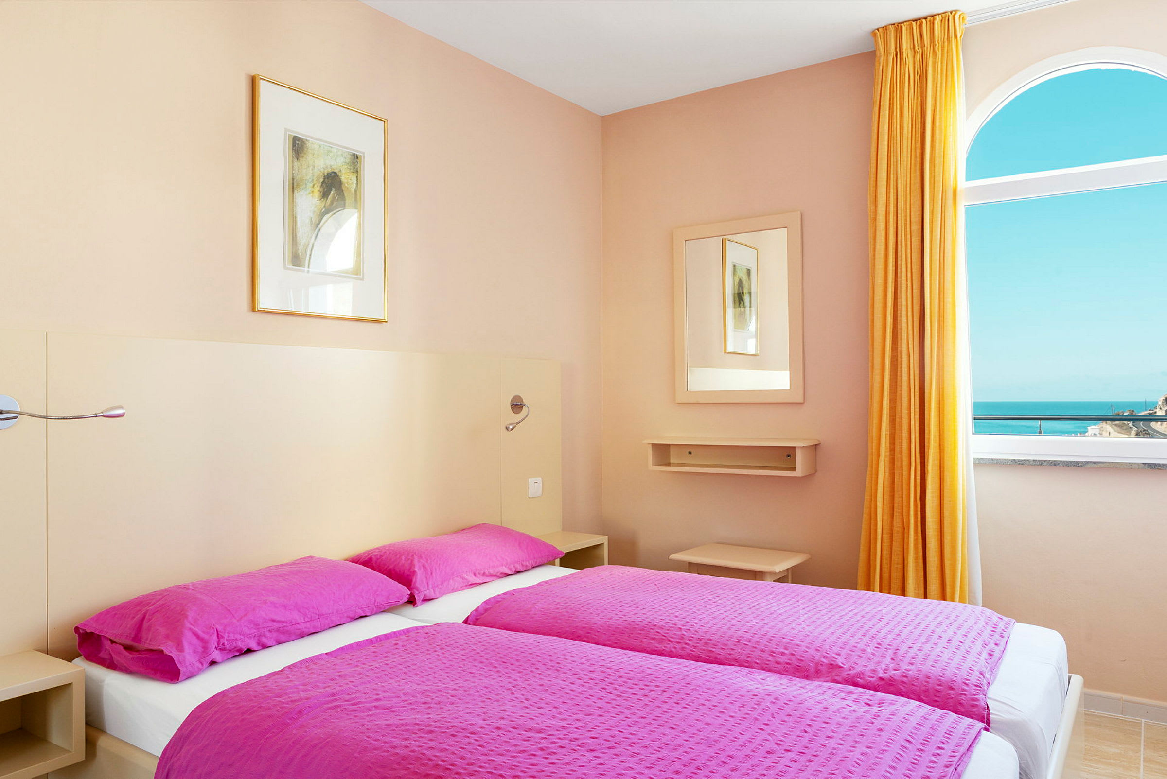 Ett hotellrum med två sängar bäddade med rosa sängkläder. Ett fönster med utsikt över havet syns till höger. (AI-genererad text)