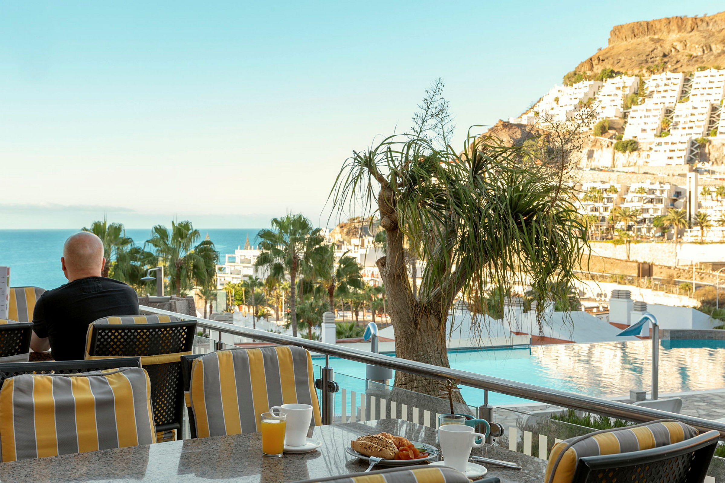 En person sitter på en terrass med utsikt över havet, träd och byggnader, med en kopp kaffe och frukost framför sig. (AI-genererad text)