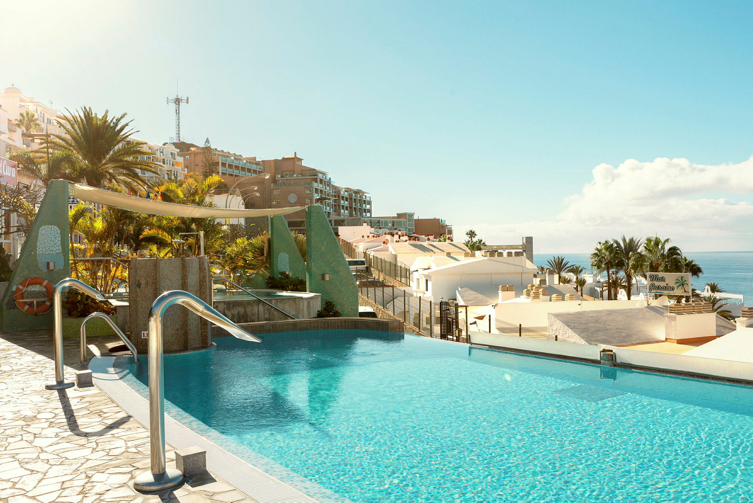En infinitypool med utsikt över hustak, palmer och havet i ett soligt landskap. (AI-genererad text)