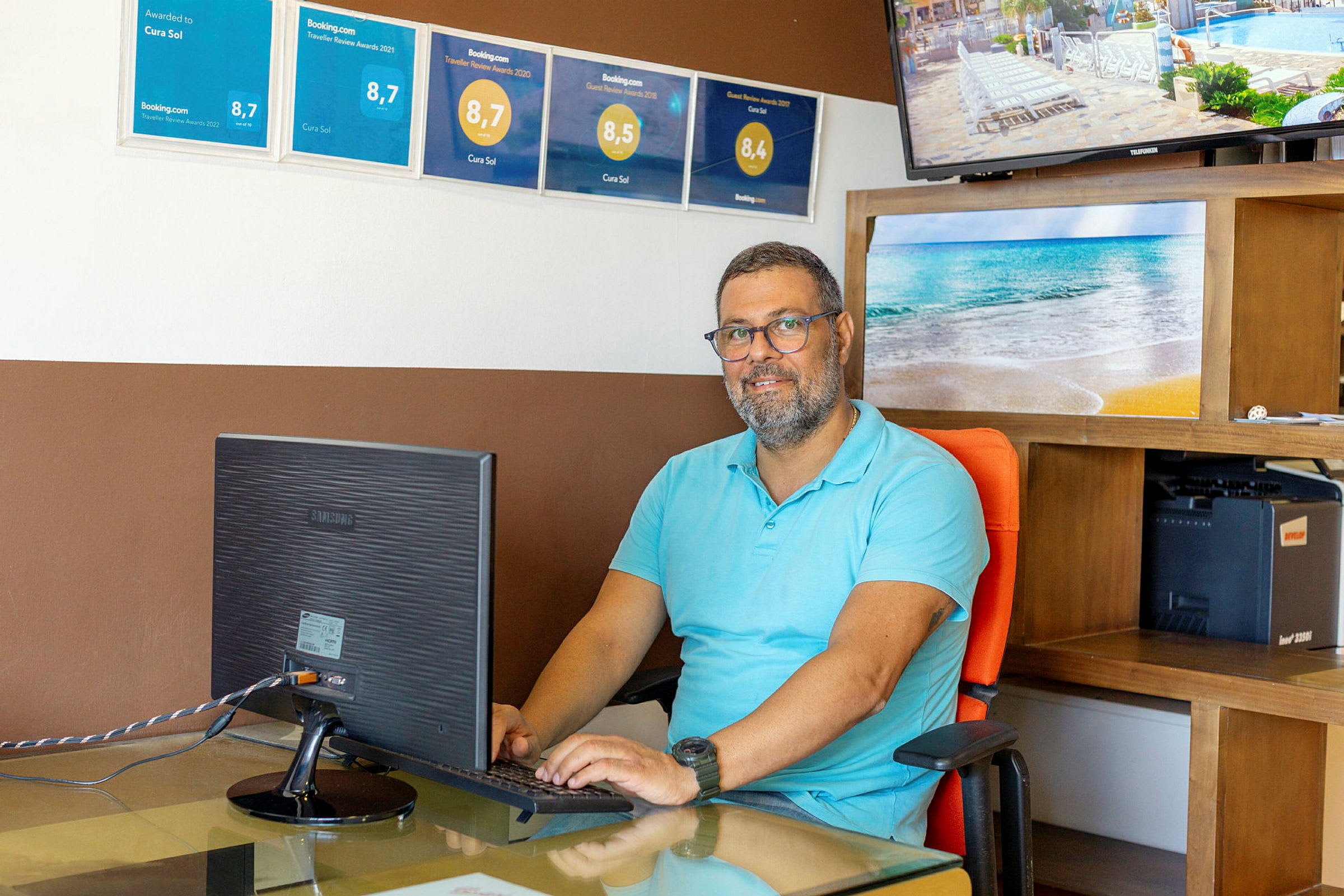 En person sitter vid ett skrivbord framför en datorskärm. På väggen bakom syns flera ramas med utmärkelser samt en TV som visar en semesteranläggning. (AI-genererad text)