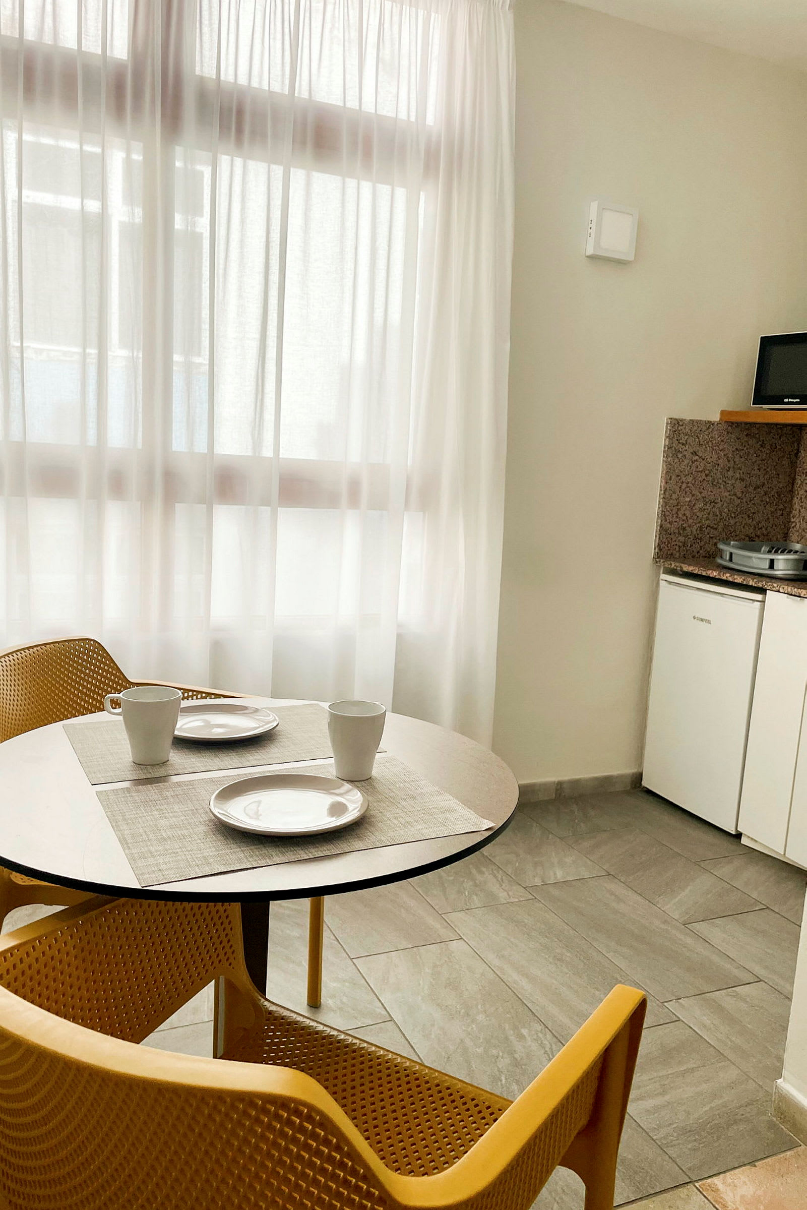 Ett matbord med två stolar, två tallrikar och koppar framför ett fönster. Köksutrustning och kylskåp syns till höger. (AI-genererad text)