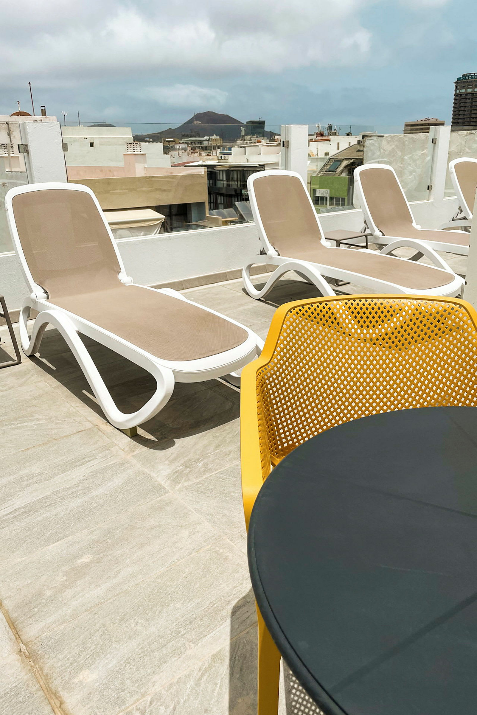 En solterrass med solstolar och ett bord med gula stolar i förgrunden, stadsvy och berg i bakgrunden. (AI-genererad text)