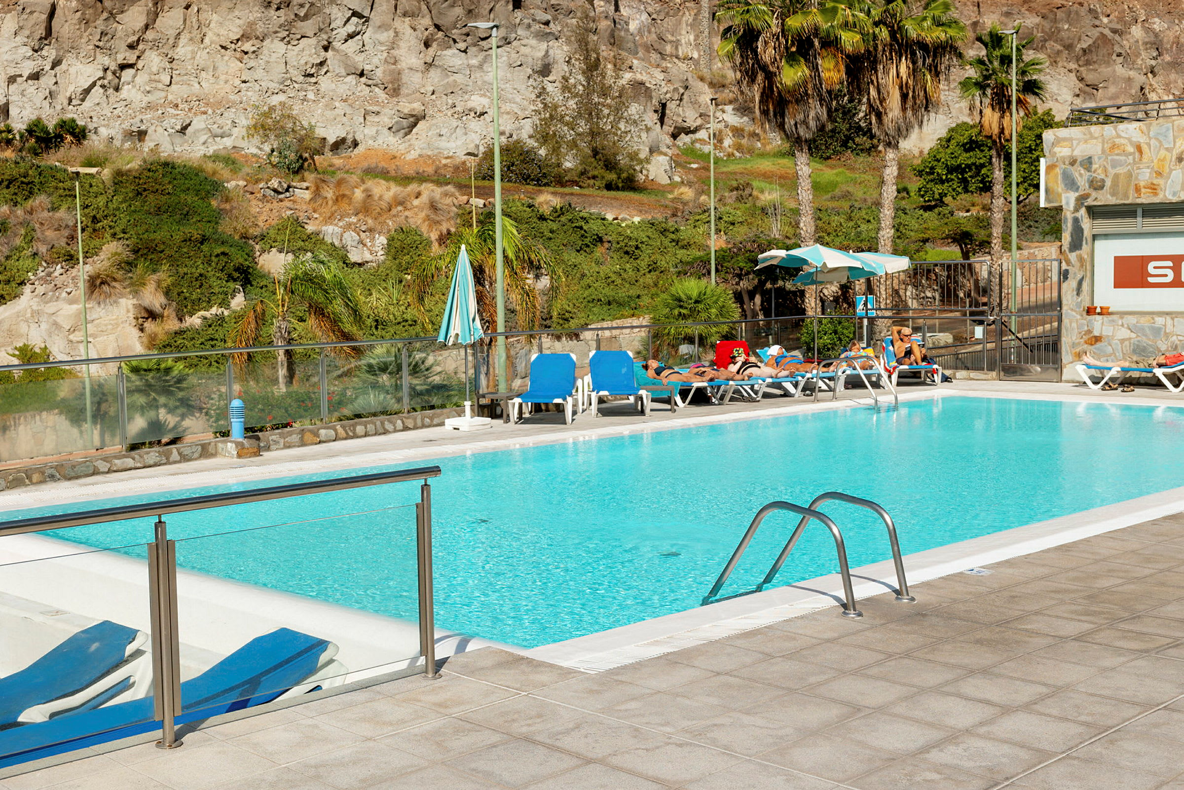 En swimmingpool omgiven av solstolar och parasoller, med klippor och växtlighet i bakgrunden. (AI-genererad text)
