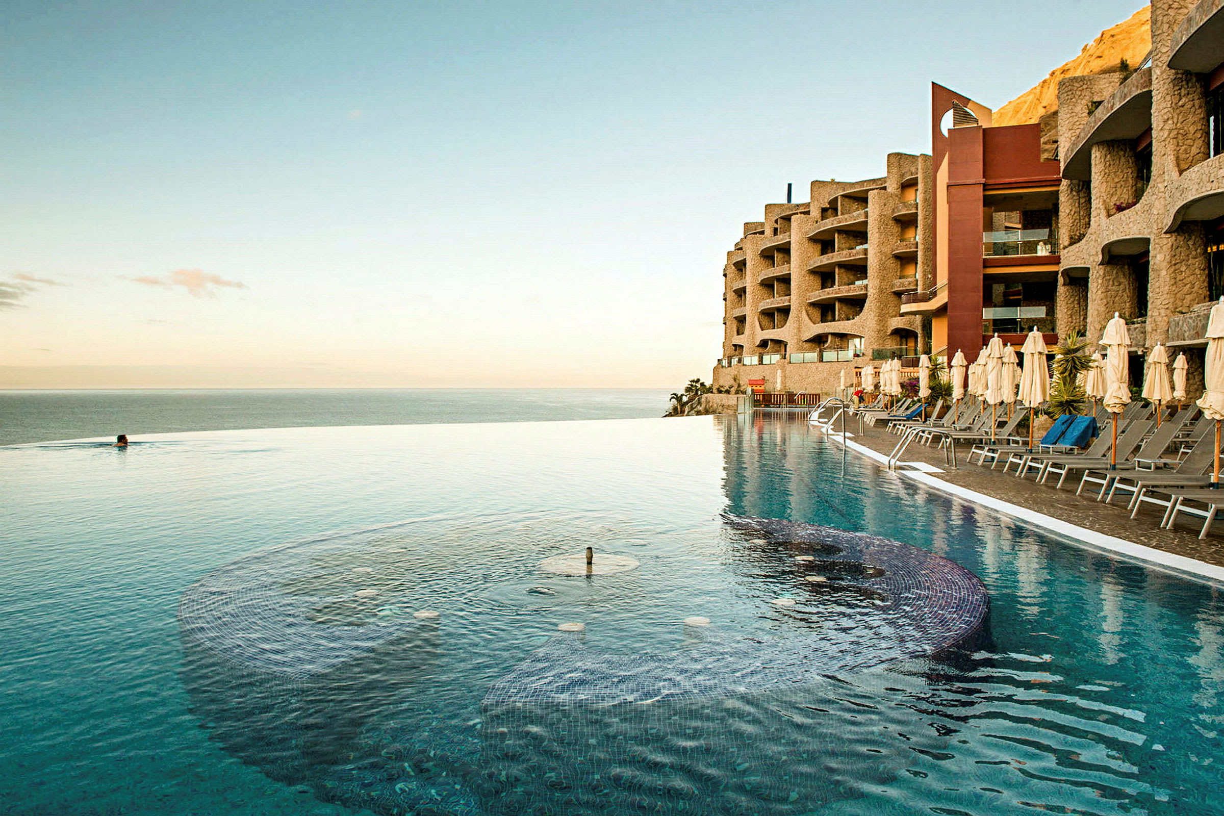 En infinitypool vid havet med solbäddar och paraplyer på sidan, samt ett hotell med modern arkitektur i bakgrunden. (AI-genererad text)