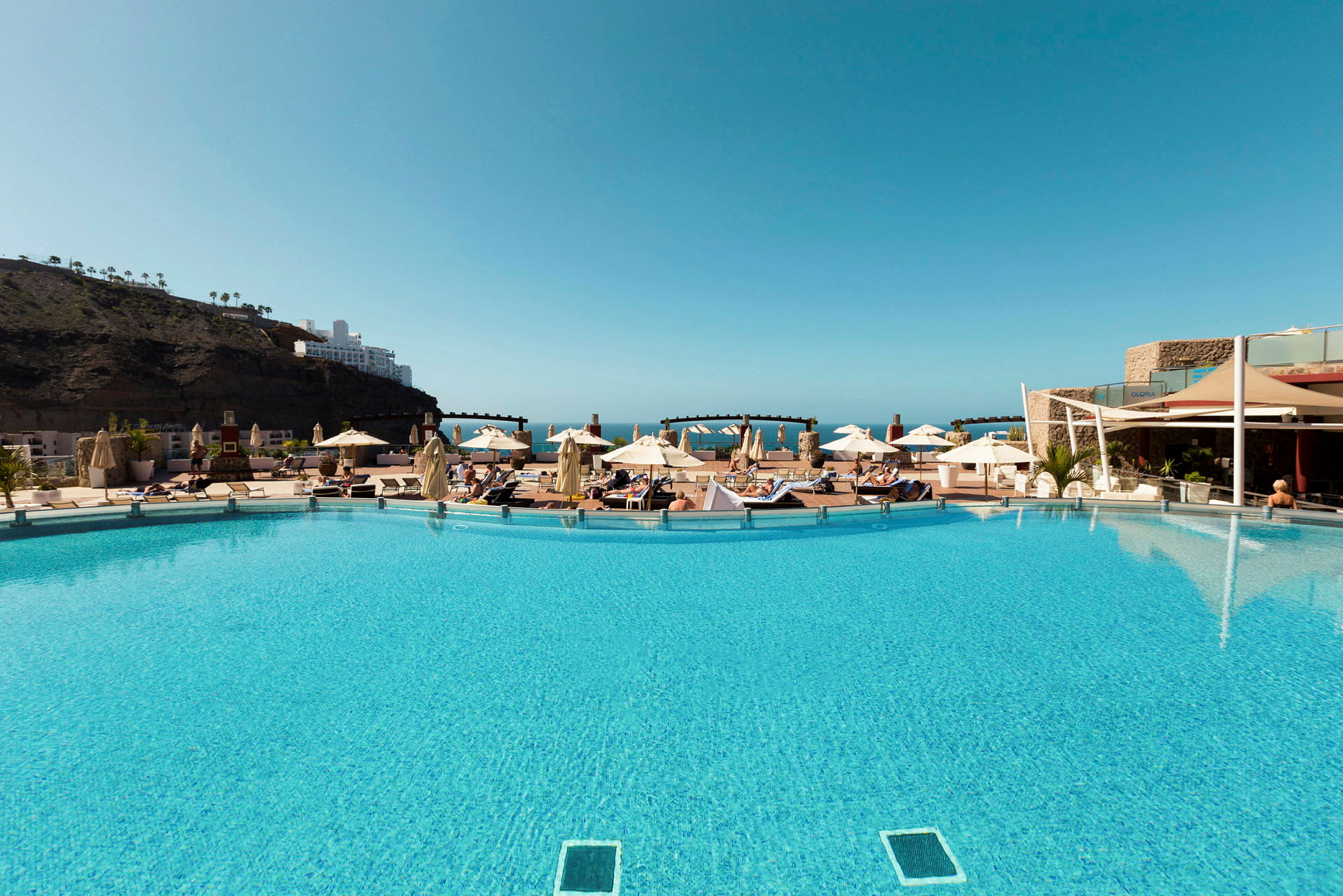 En swimmingpool med solstolar och parasoller, omgiven av byggnader och berg under en klarblå himmel. (AI-genererad text)