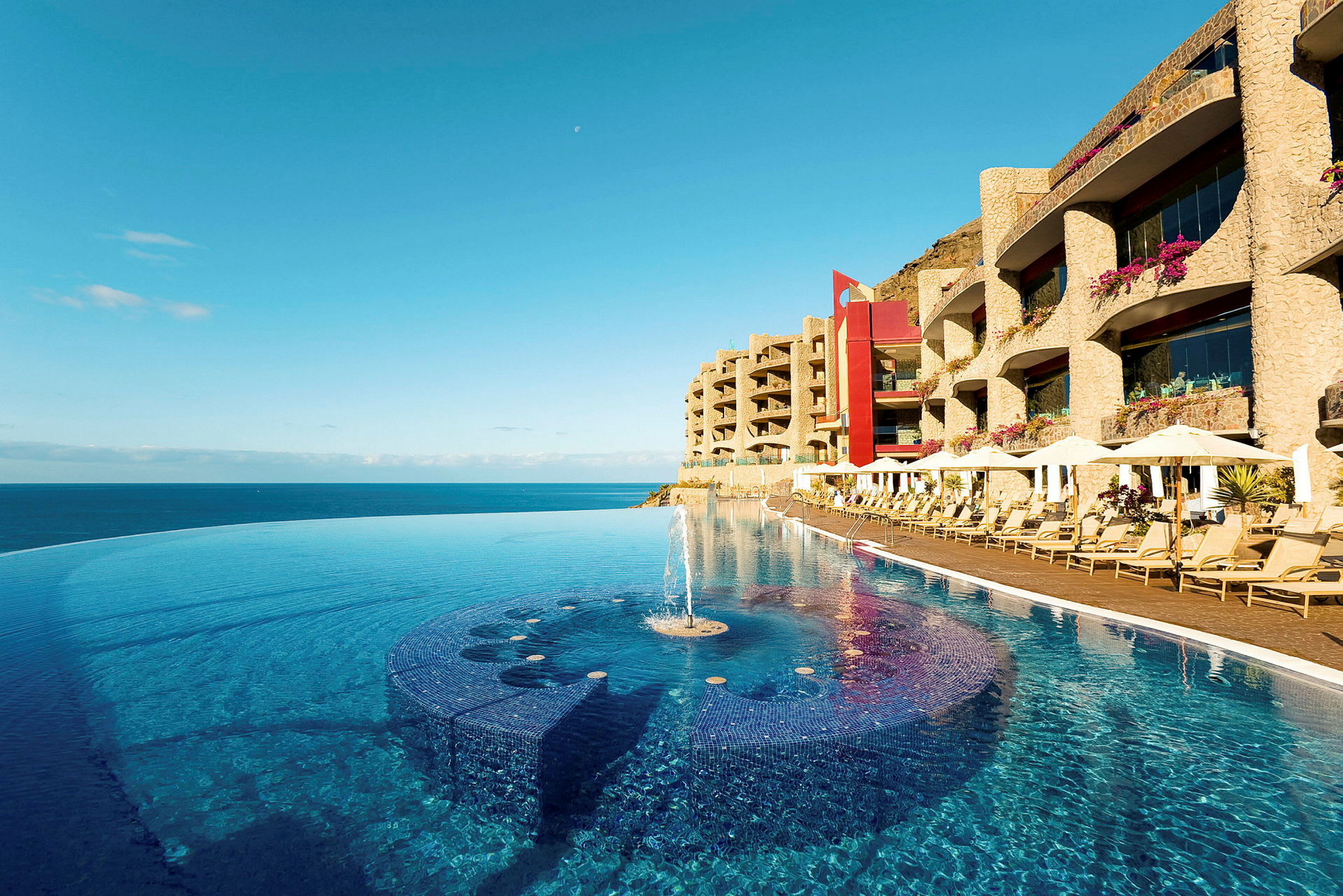 En infinitypool vid strandkanten med solstolar och hotellbyggnader i bakgrunden. (AI-genererad text)