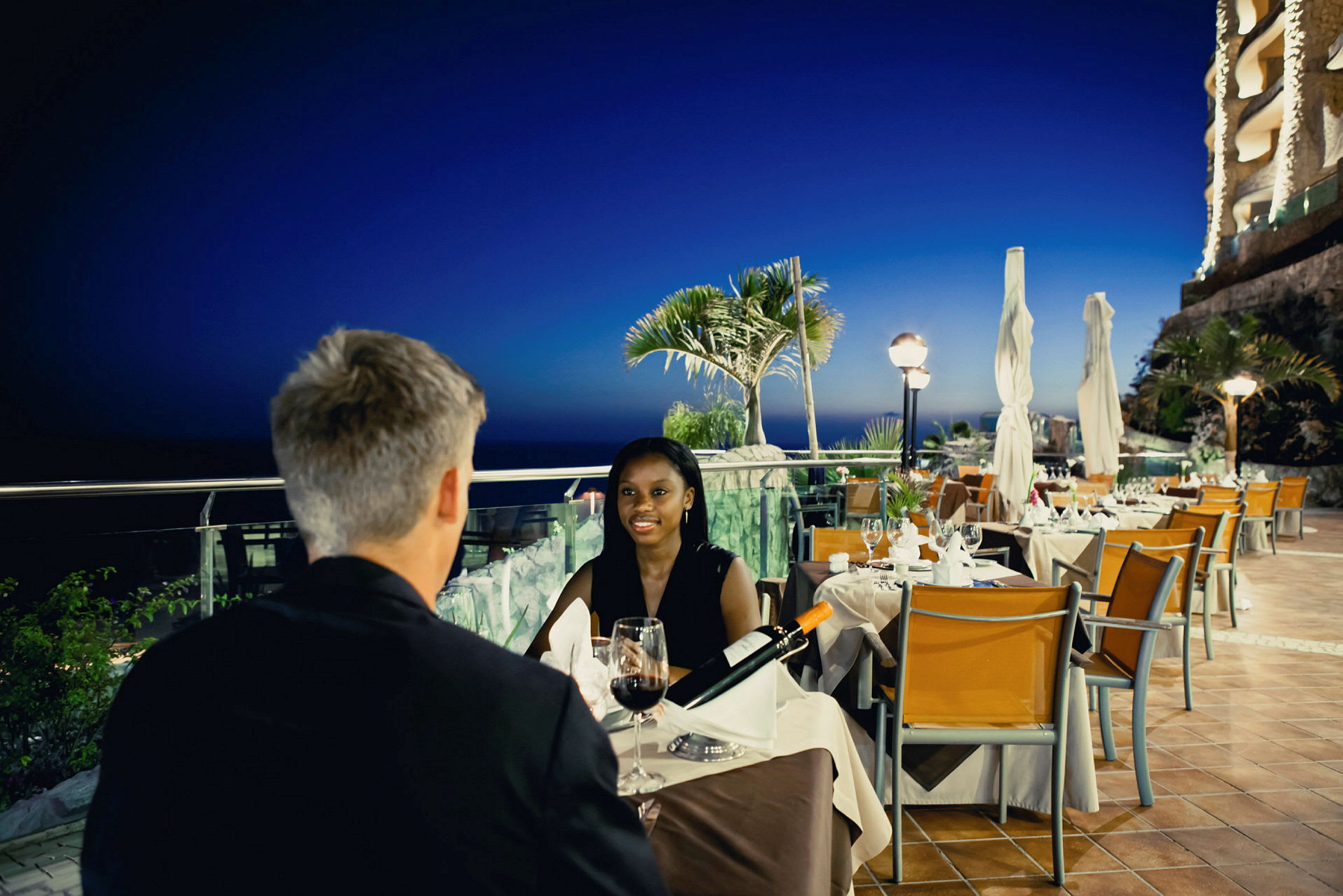 Två personer vid ett bord på en restaurang med kvällsutsikt över havet och tropiska växter. (AI-genererad text)
