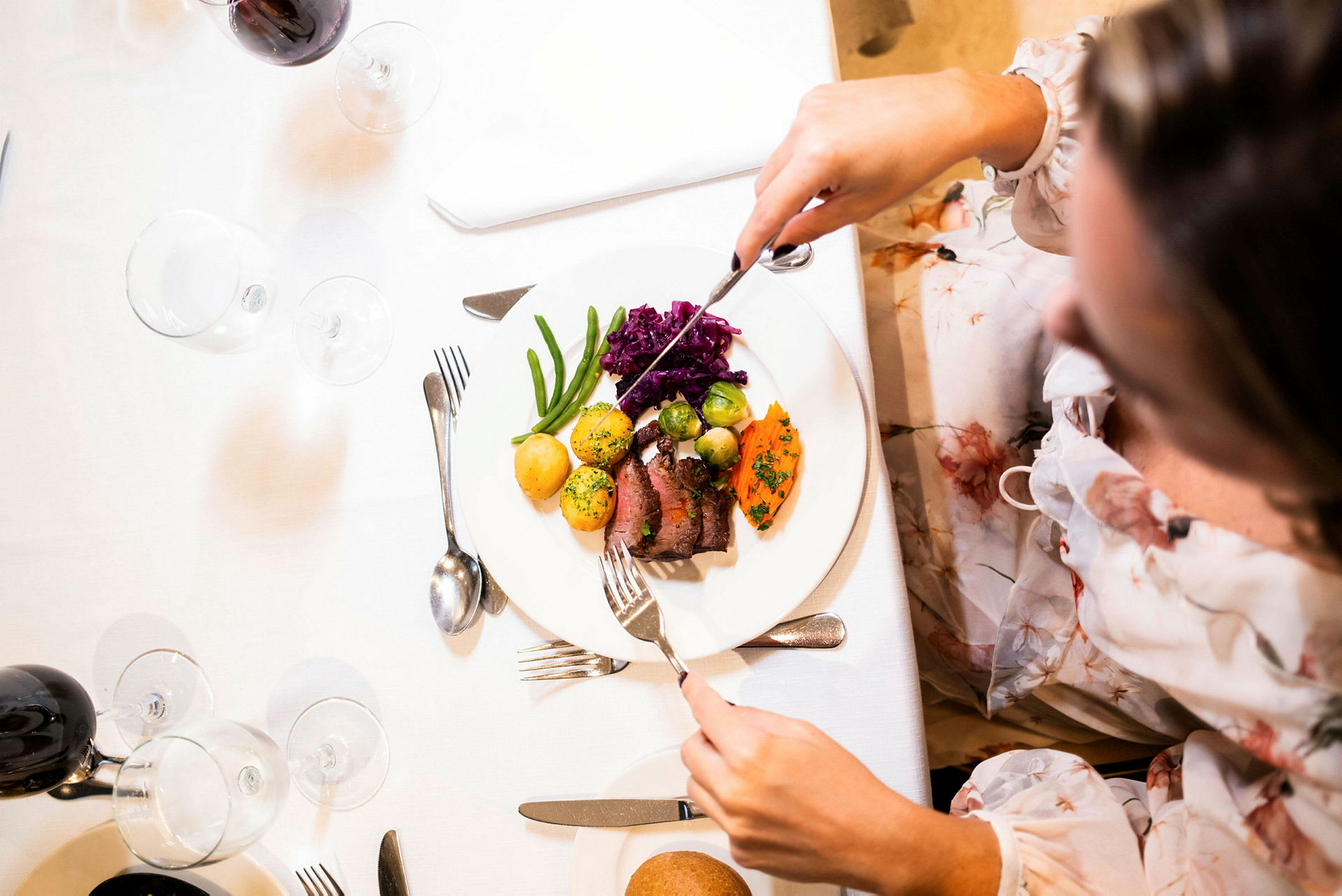 En tallrik med en portion mat innehållande kött, grönsaker och potatis ligger på en vit duk. En person håller en kniv och gaffel. (AI-genererad text)
