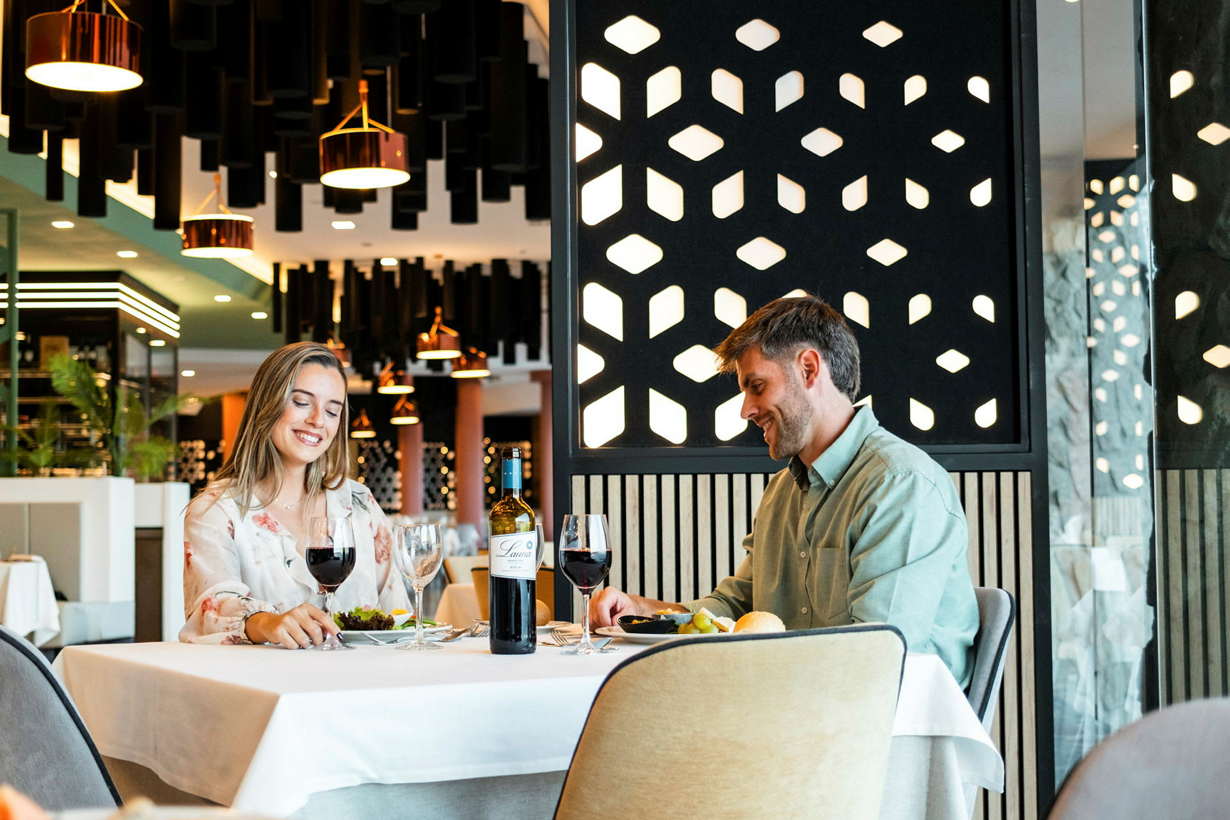 Två personer sitter vid ett bord i en restaurang med modern inredning. På bordet finns glas, ett vinflaska och tallrikar med mat. (AI-genererad text)