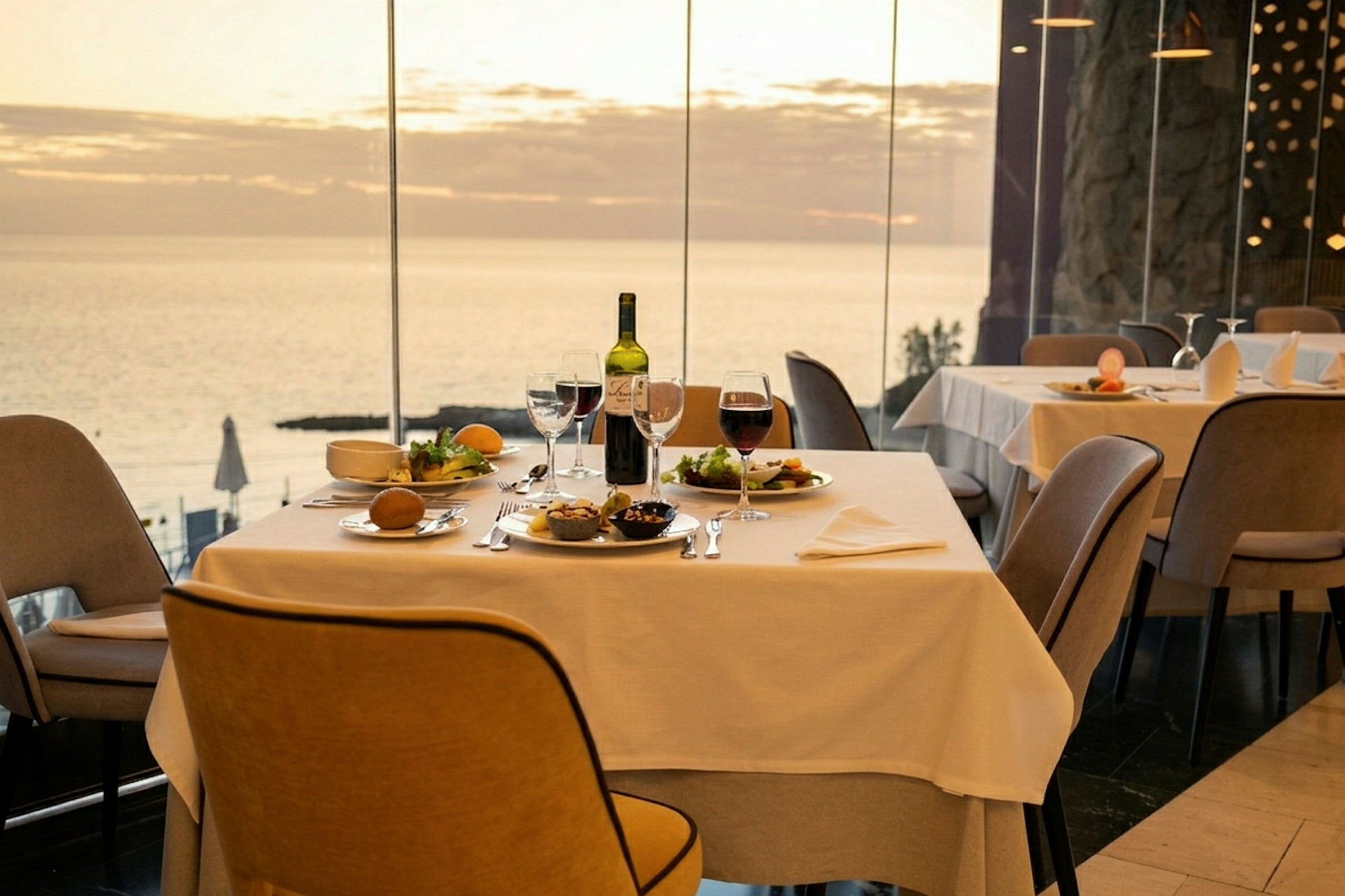 Ett bord i en restaurang med vita dukar, matuppsättning och vin på bordet, med havsutsikt genom stora fönster. (AI-genererad text)