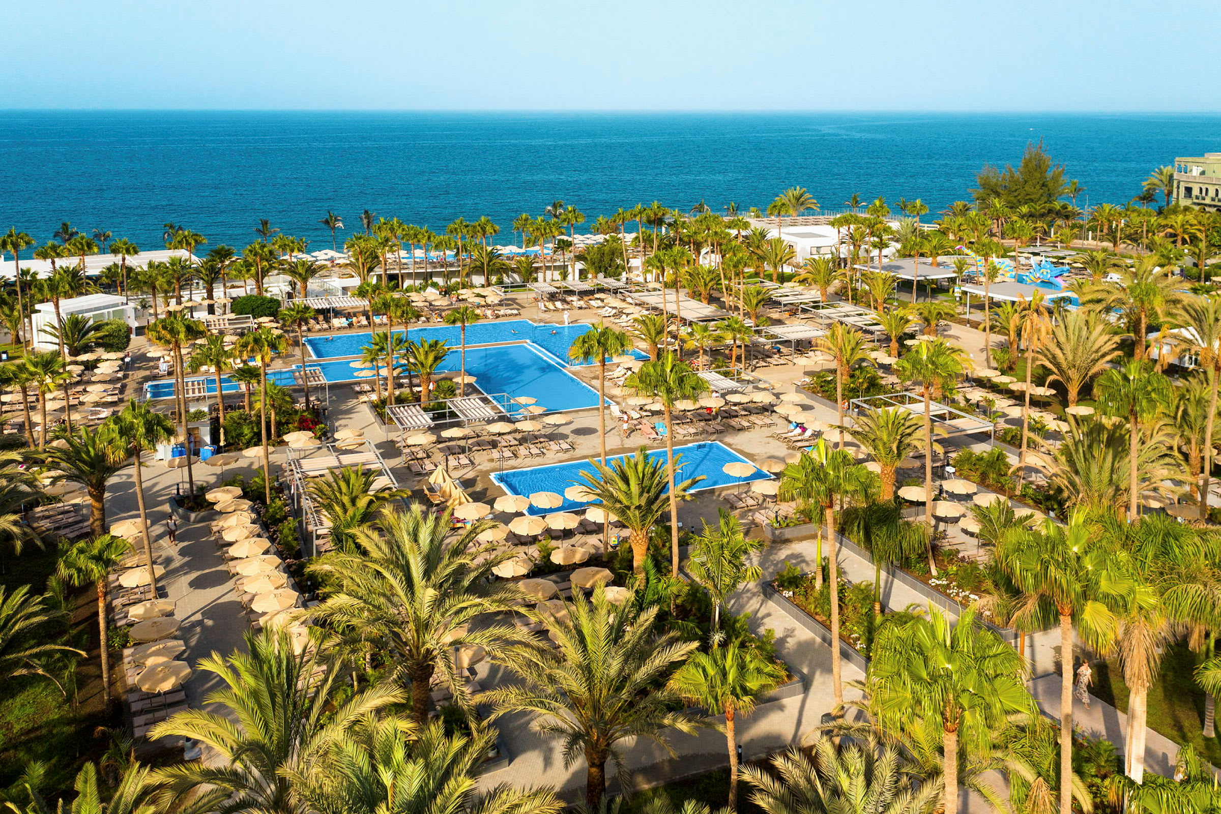Flygbild över en hotellresort med pooler och palmer, med havet i bakgrunden. (AI-genererad text)