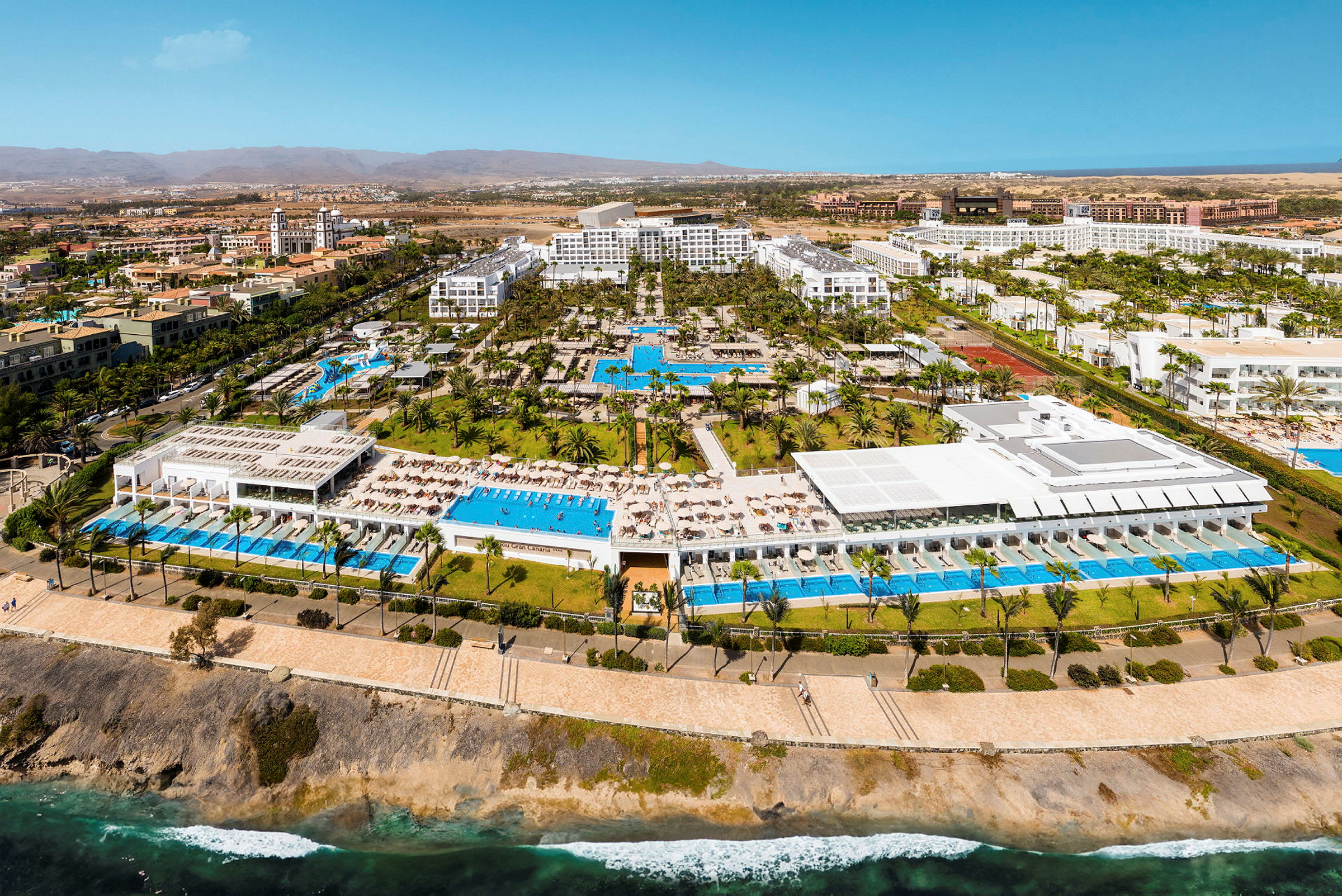 Flygbild av en resort med pooler, byggnader och träd vid kusten. (AI-genererad text)
