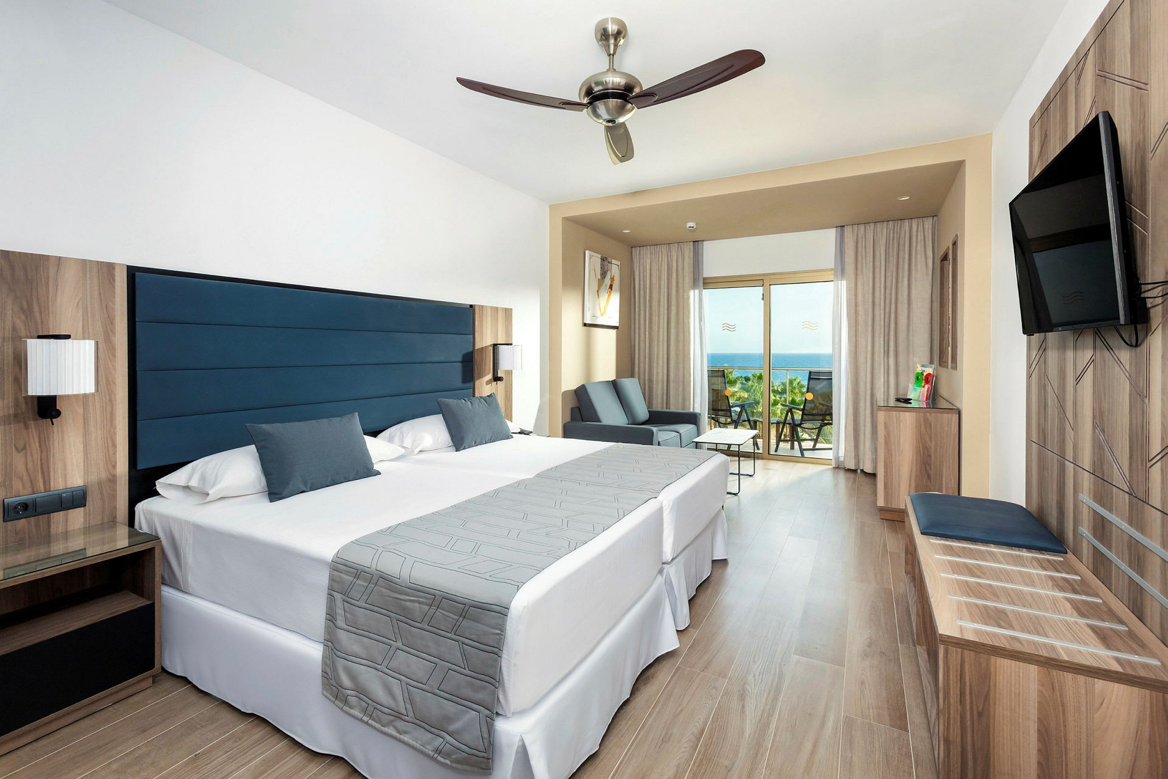 Ett hotellrum med två sängar, ett sittområde och en balkong som har utsikt över havet. (AI-genererad text)