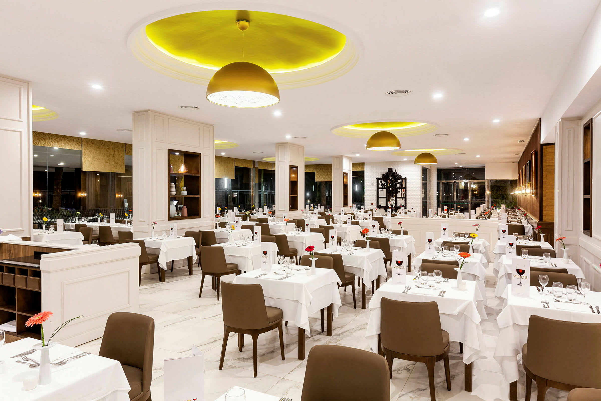 En interiörbild av en ljus och elegant restaurang med många bord uppdukade med vita dukar, bruna stolar och dekorationer. (AI-genererad text)