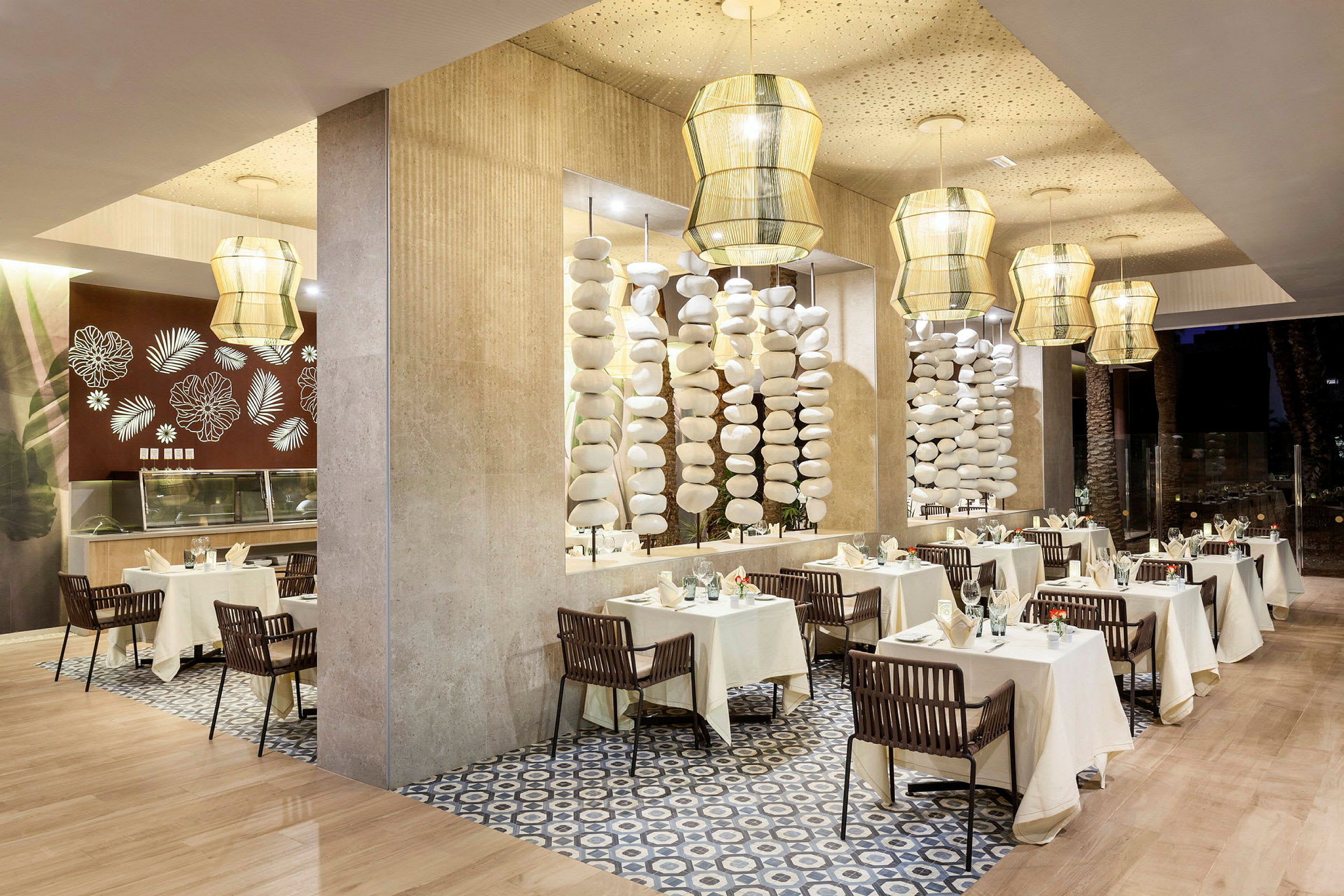 En elegant restauranginteriör med bord täckta av vita dukar och stilfullt dekorerade med porslin och glas. Ljusa hängande lampor och dekorativa pelare kompletterar designen. (AI-genererad text)