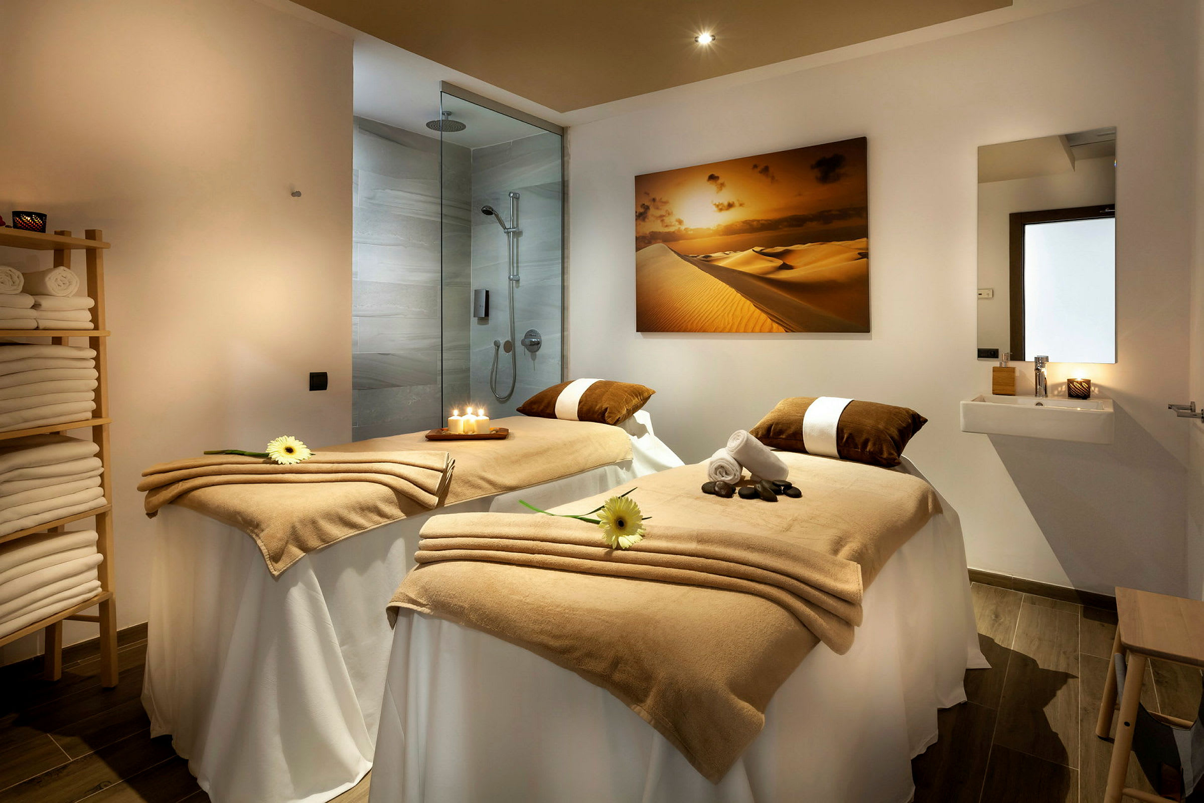 Ett spa-behandlingsrum med två massagebänkar täckta med beige filtar, ljus och dekorativa blommor. En dusch och handfat syns i bakgrunden. (AI-genererad text)
