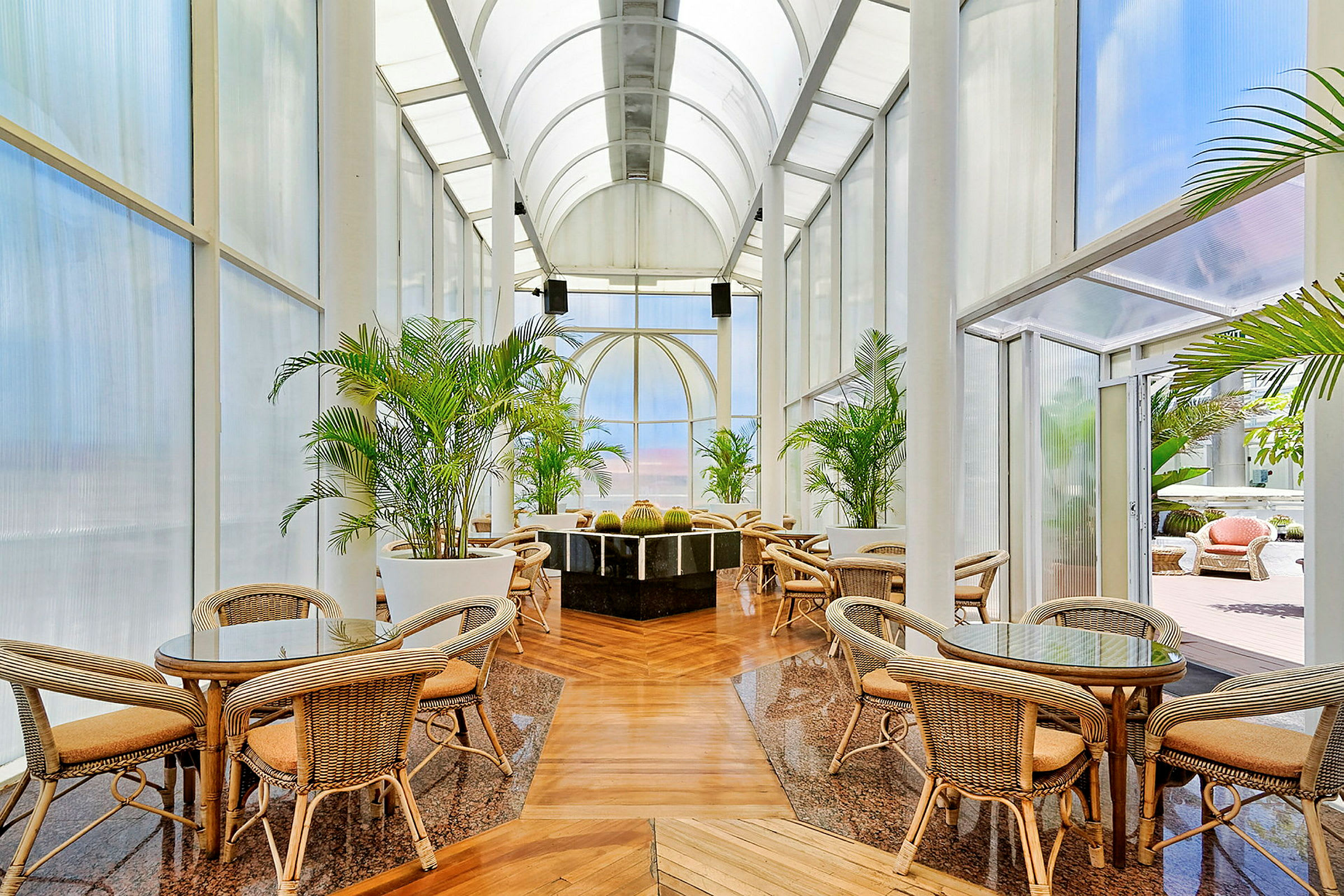 Rostmöbler och bord under högt valvat tak med stora glaspartier omgiven av gröna växter. (AI-genererad text)