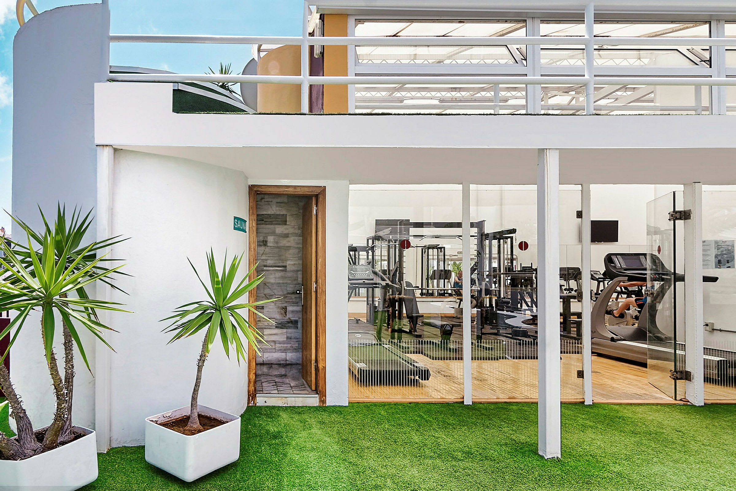 Exteriör av ett gym med glasväggar och en bastudörr, samt gröna planteringar i krukor framför. (AI-genererad text)
