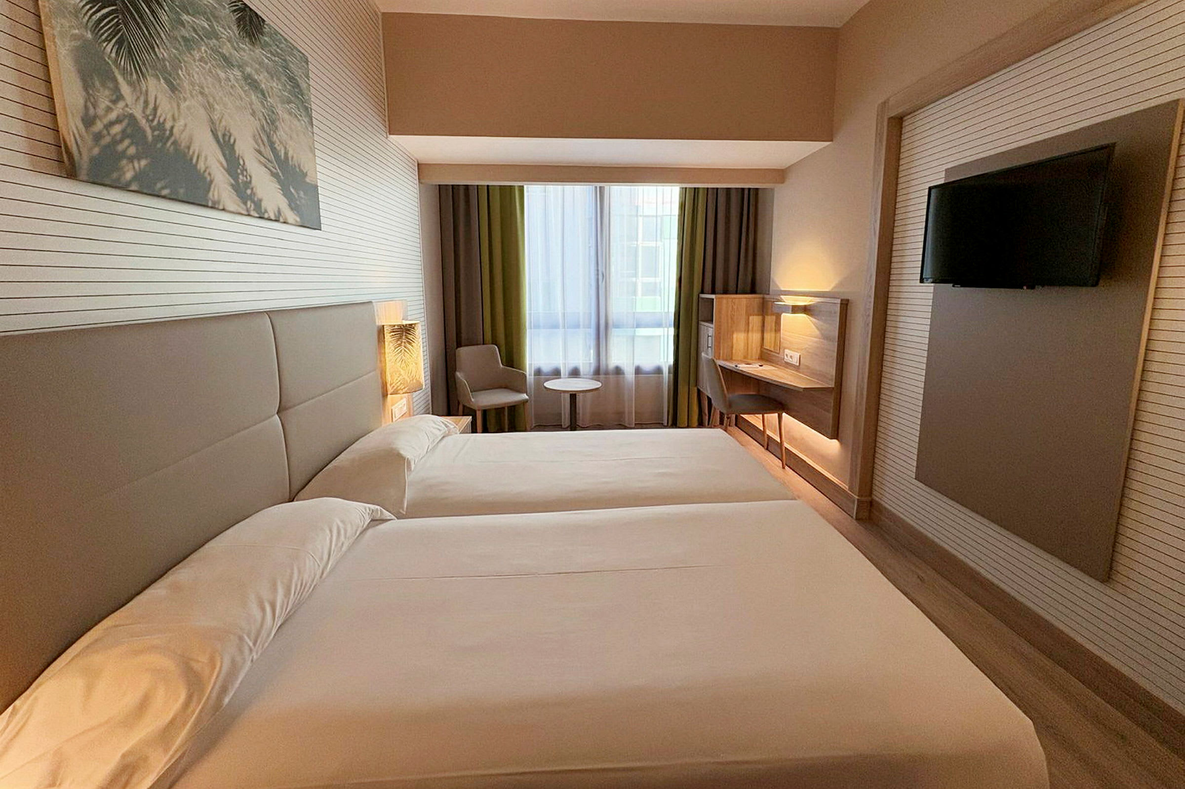 Hotelrum med två enkelsängar, ett fönster med gardiner, sittplats med bord och en monterad platt-TV. (AI-genererad text)