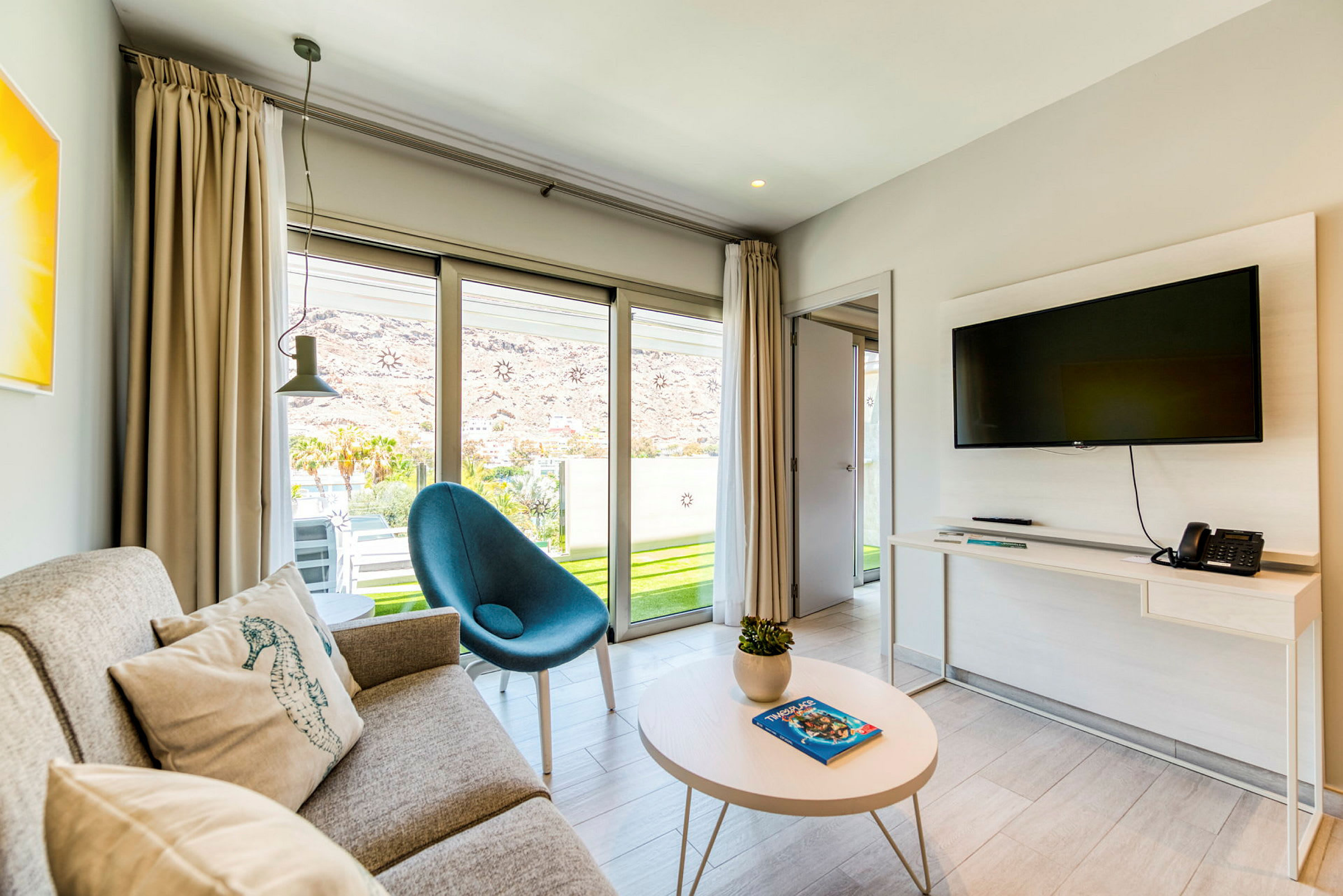 Ett modernt hotellrum med soffa, fåtölj och väggmonterad TV, samt glasdörrar som leder ut till en terrass. (AI-genererad text)