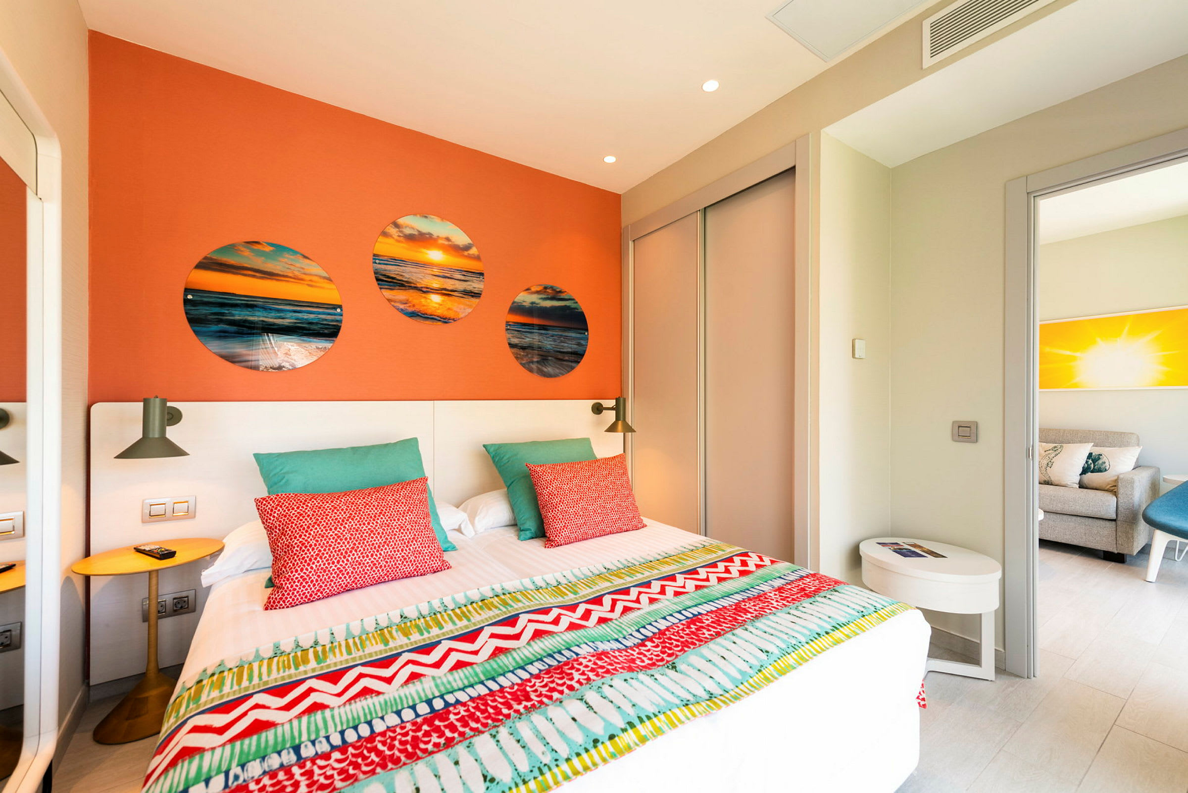 Ett hotellrum med orange vägg, säng med färgglad överkast och kuddar samt inredningsdetaljer. (AI-genererad text)