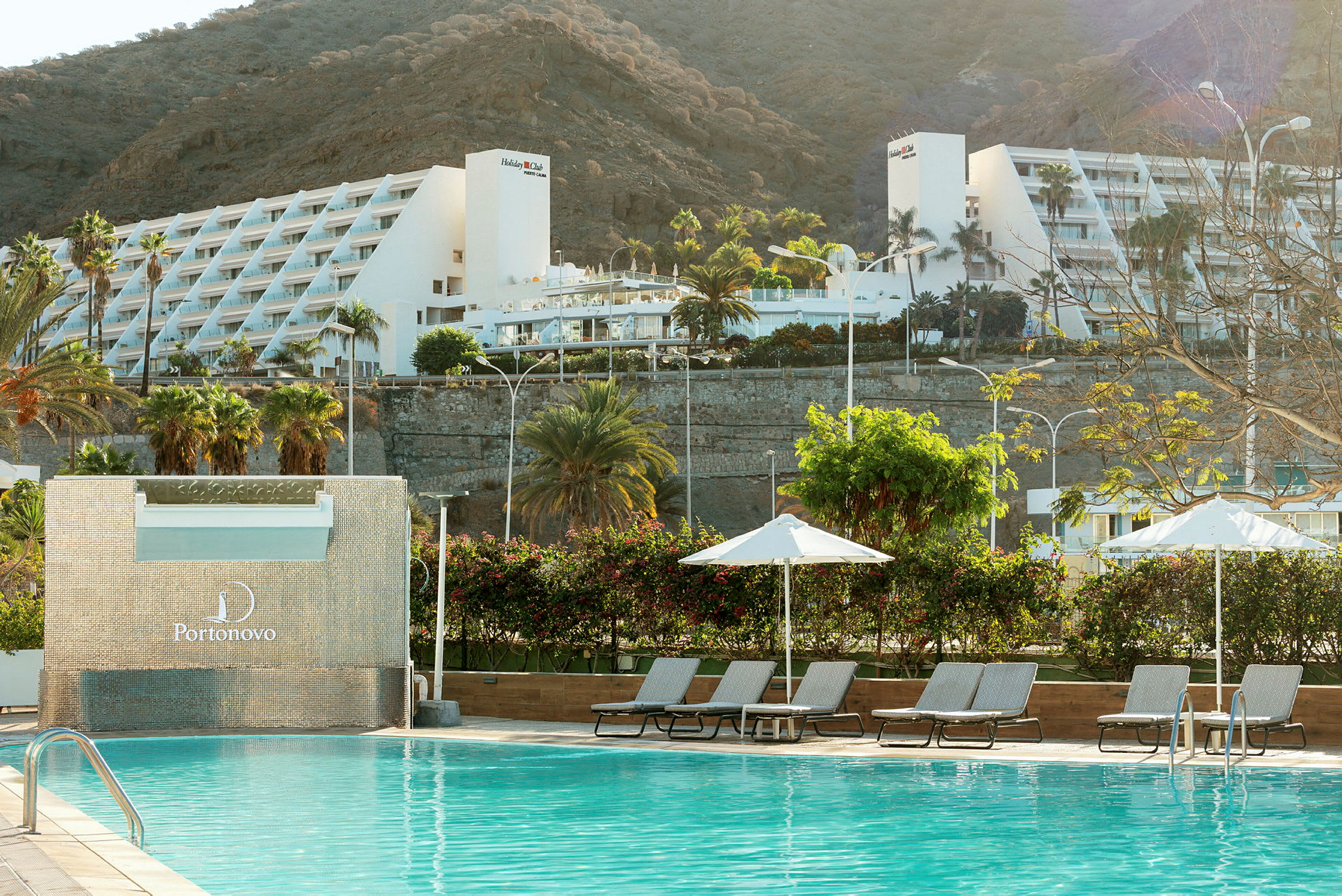 En pool med solstolar och parasoller, med en vit semesteranläggning och berg i bakgrunden. (AI-genererad text)