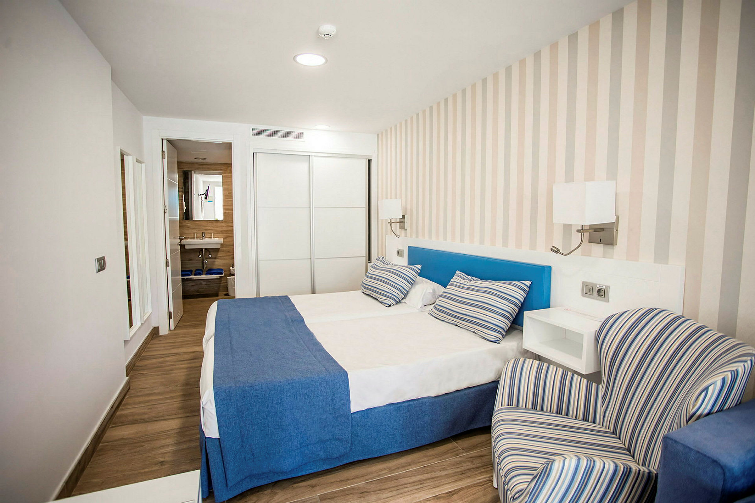 Hotellrum med en dubbelsäng, blå sängöverkast och randiga kuddar, en randig fåtölj samt synbart badrum i bakgrunden. (AI-genererad text)