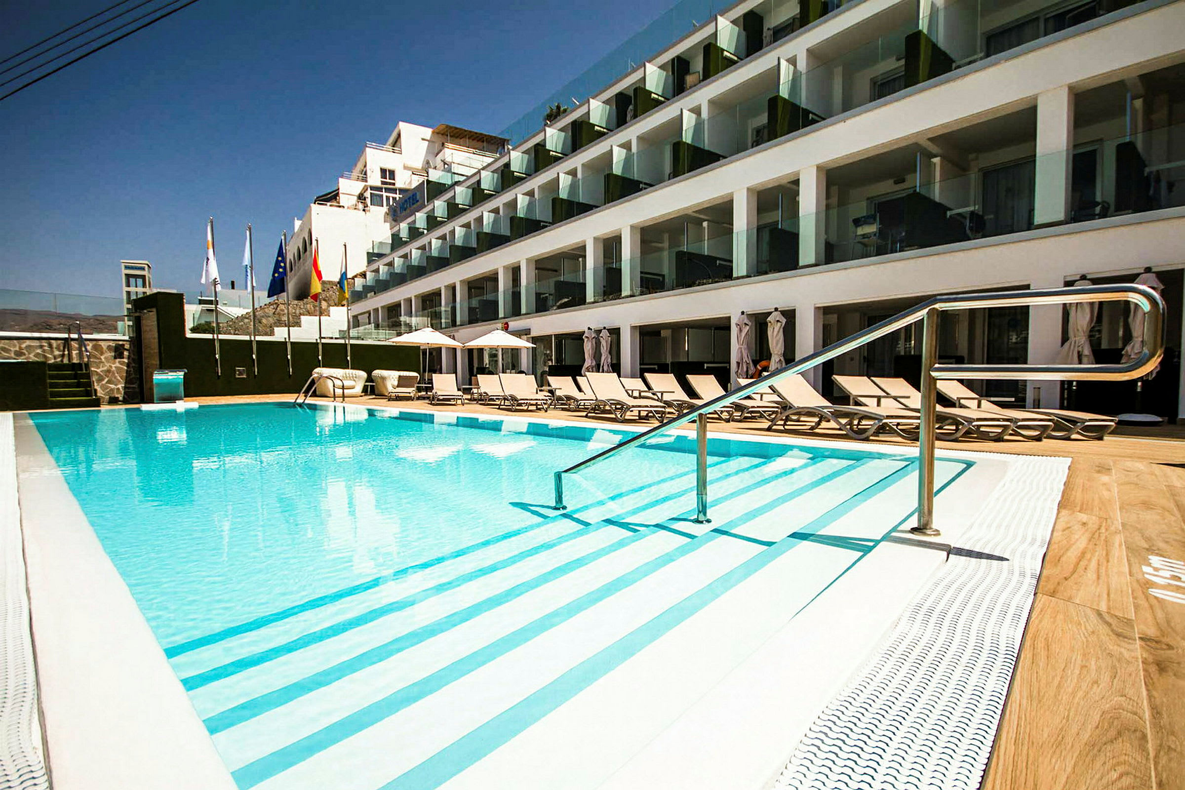Utomhuspool vid hotell med solstolar och glasbalkonger. (AI-genererad text)