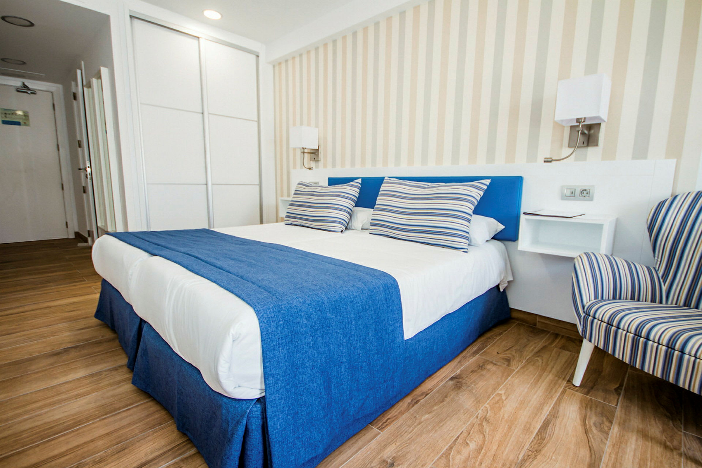En smakfull hotellrum med en dubbelsäng täckt av blå och vit sängkläder samt randiga kuddar, vid sidan av ett randigt fåtölj. (AI-genererad text)
