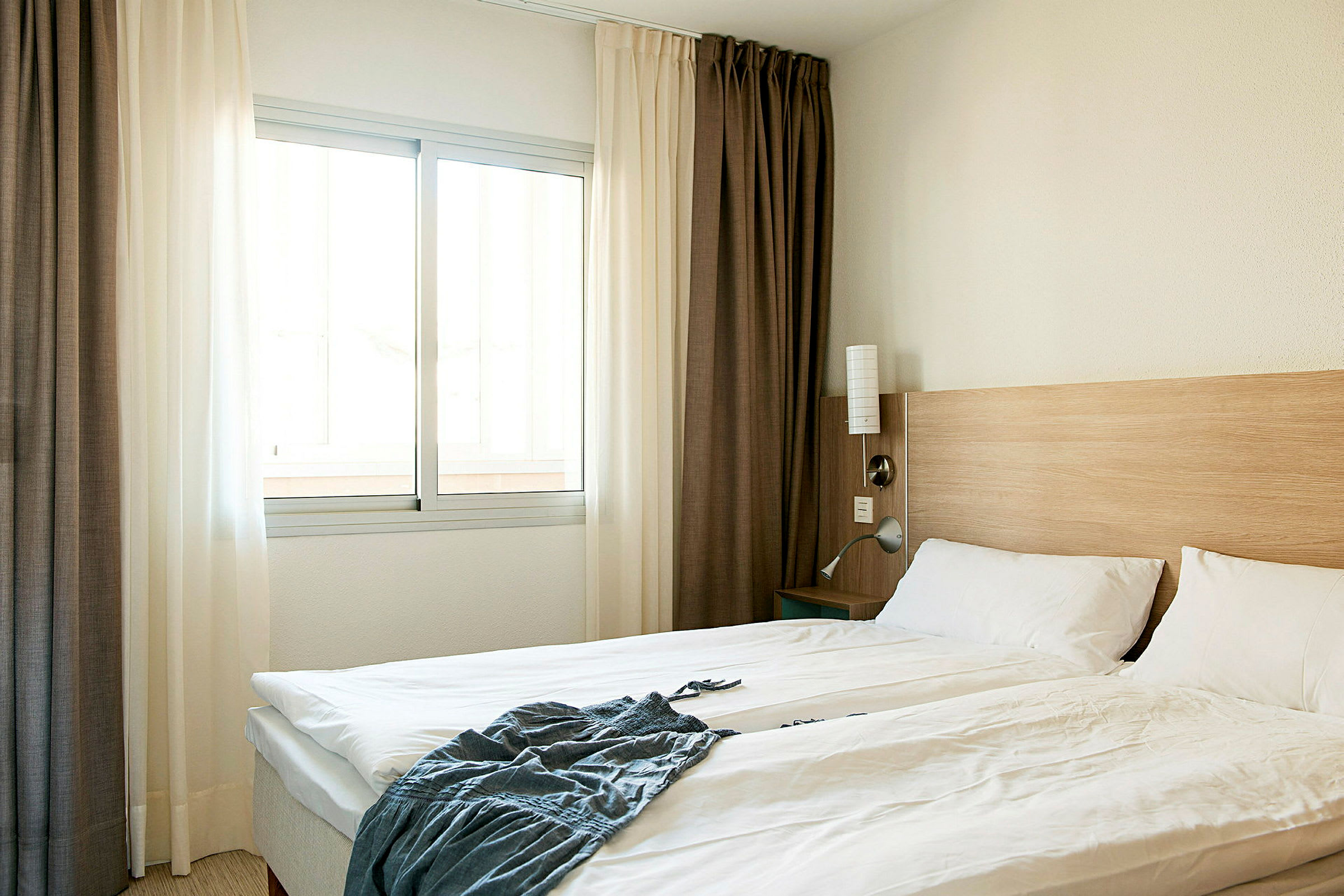 Ett hotellrum med en dubbelsäng, vita sängkläder, en trävägg vid sänggaveln och ett fönster med ljusa gardiner. (AI-genererad text)