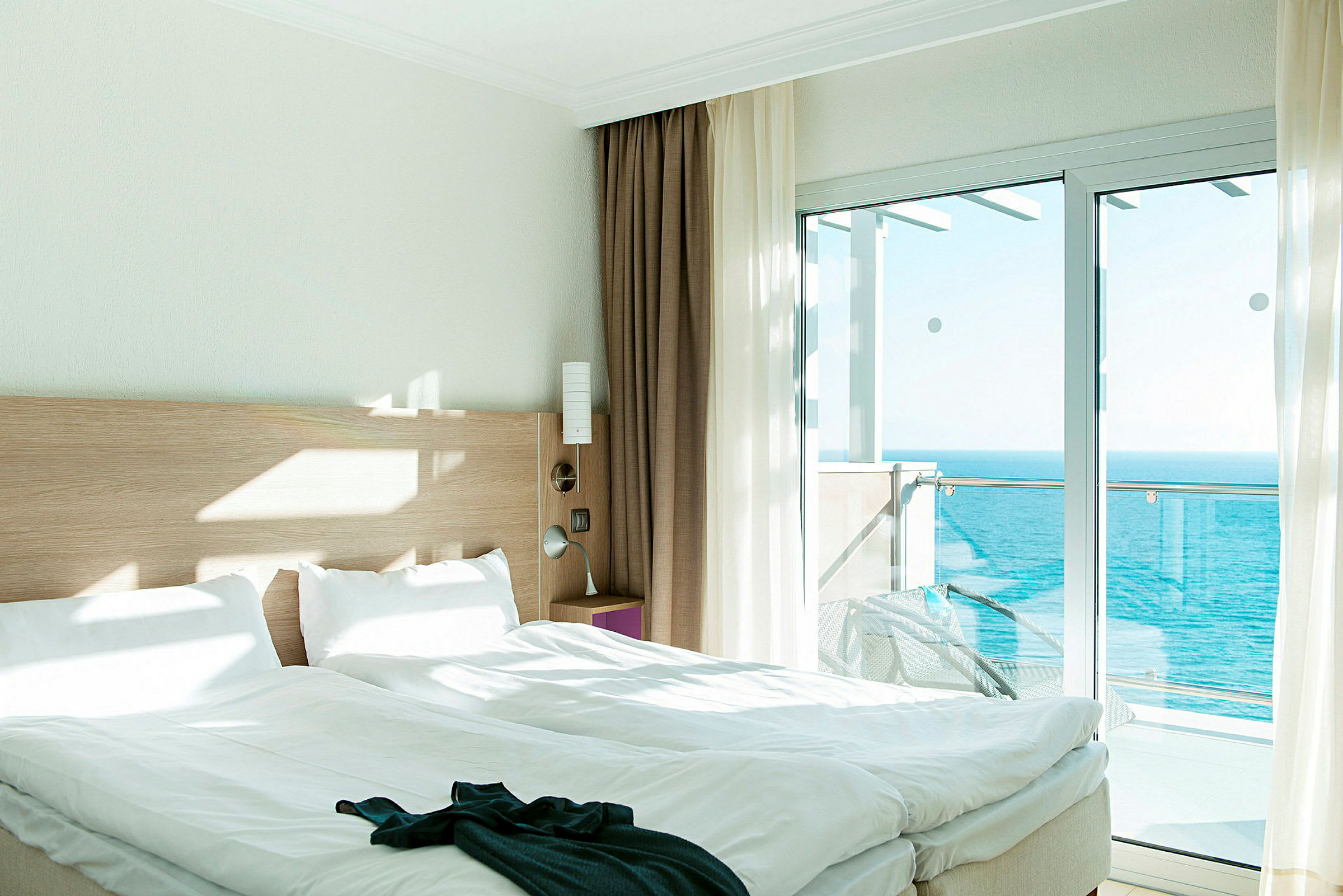 Ett hotellrum med havsutsikt. Sängen är bäddad och solens ljus lyser upp rummet genom ett glasfönster och balkong. (AI-genererad text)
