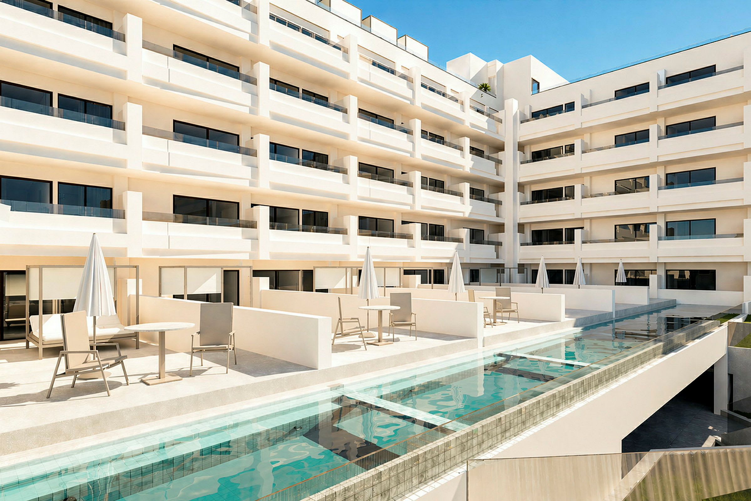 Vy över en modern hotellbyggnad med flera våningar och balkonger. Framför byggnaden finns en terrass med bord, stolar och parasoller samt en swimmingpool. (AI-genererad text)