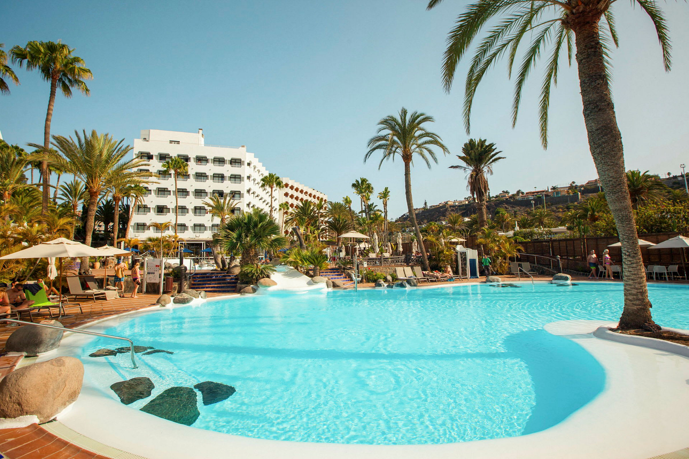 En swimmingpool omgiven av palmträd och ett hotell i bakgrunden. (AI-genererad text)