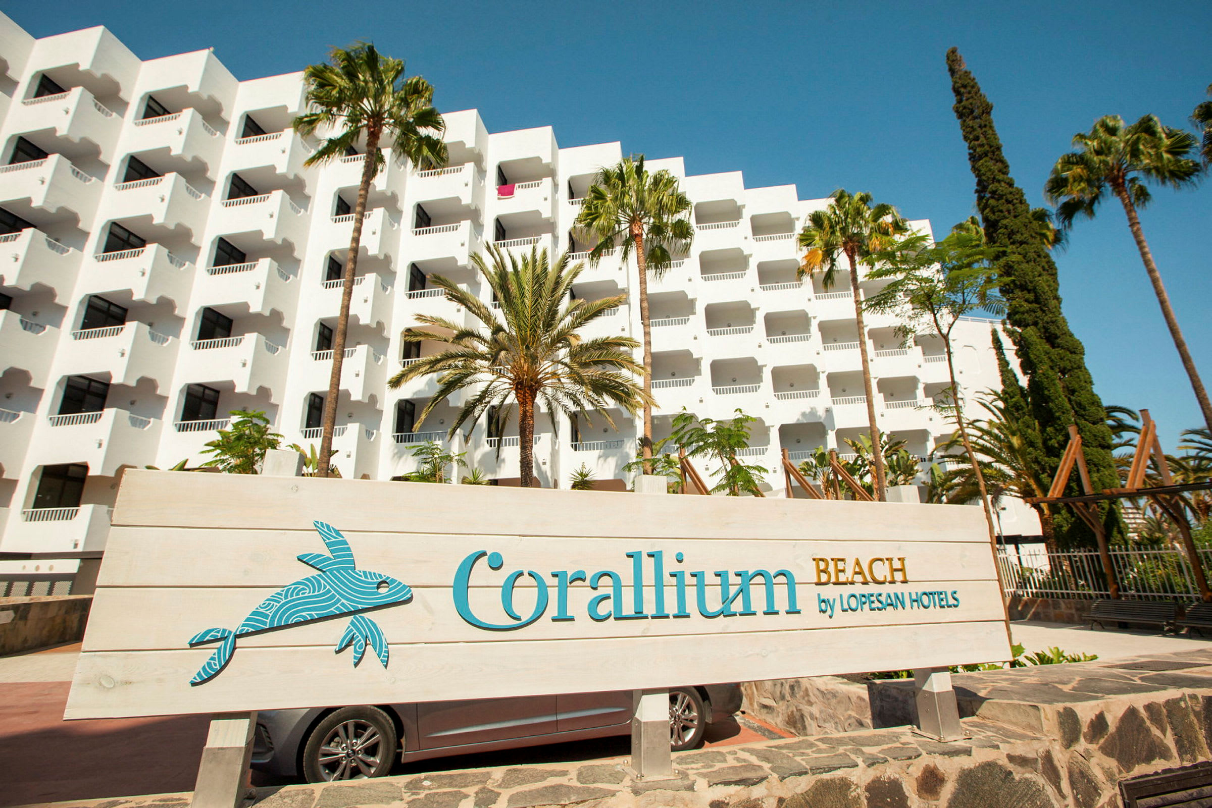 Vit hotellbyggnad med balkonger, omgiven av palmträd, samt en skylt som visar texten 'Corallium Beach by Lopesan Hotels'. (AI-genererad text)