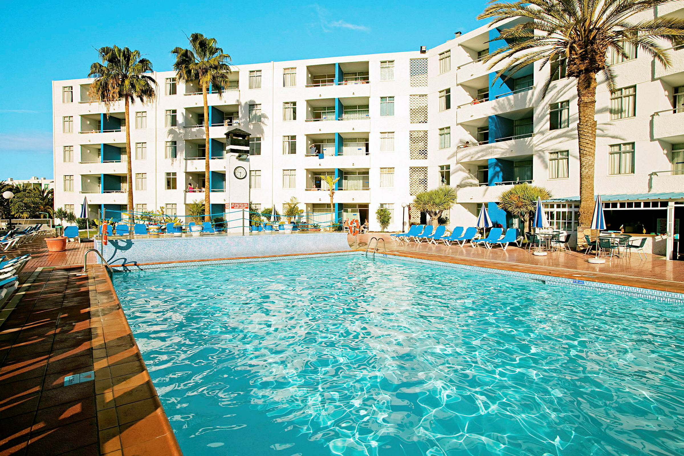 En pool vid ett hotell med solstolar, parasoller och palmer i bakgrunden. (AI-genererad text)