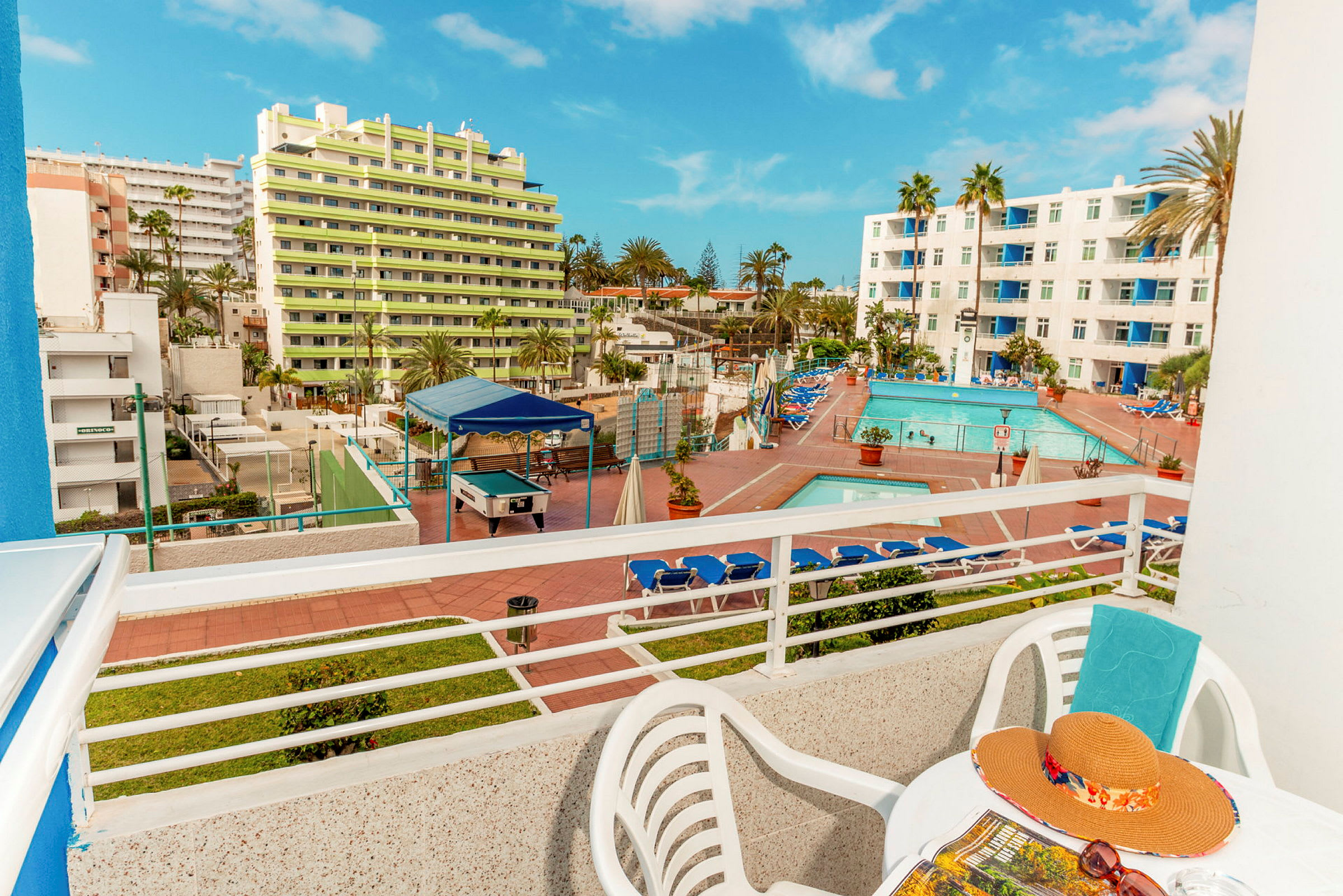 Utsikt från en balkong som visar ett hotellområde med pool och solstolar samt byggnader i bakgrunden. (AI-genererad text)