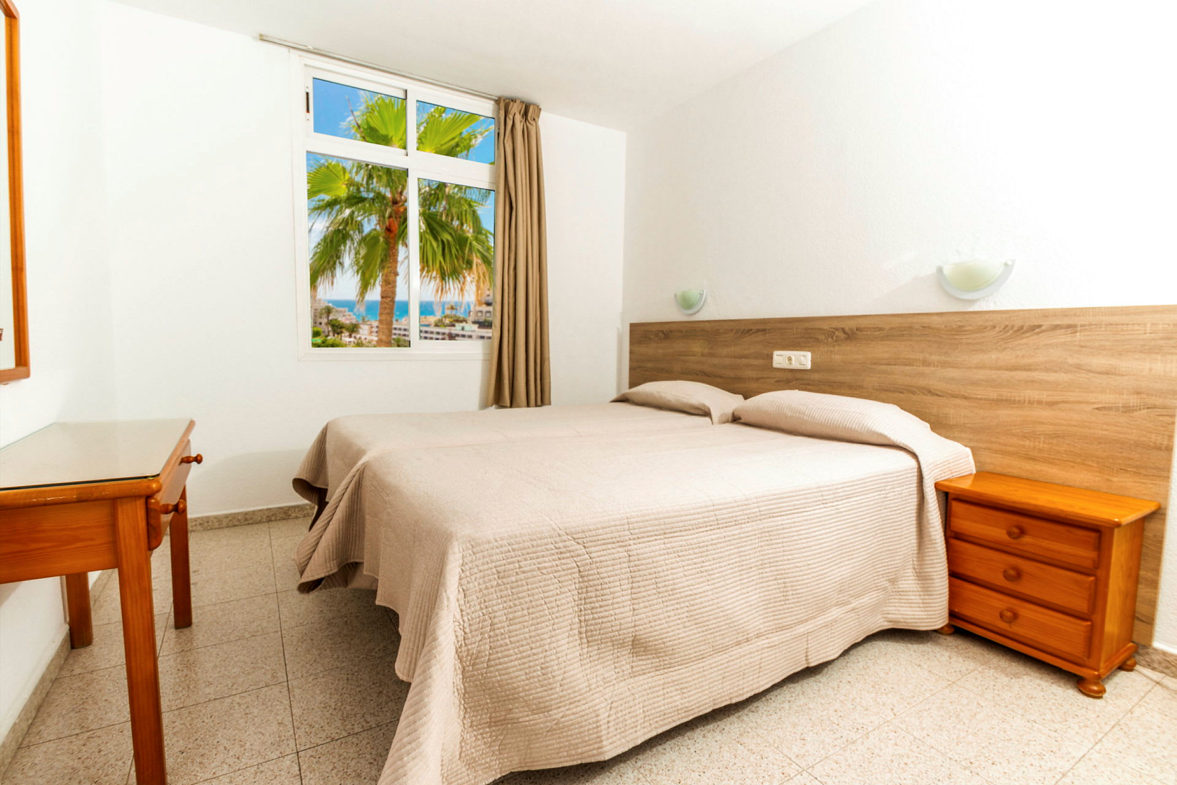 Ett hotellrum med två enkelsängar klädda i beige sängkläder. En vägg har ett fönster som vetter mot en palm och havet. (AI-genererad text)