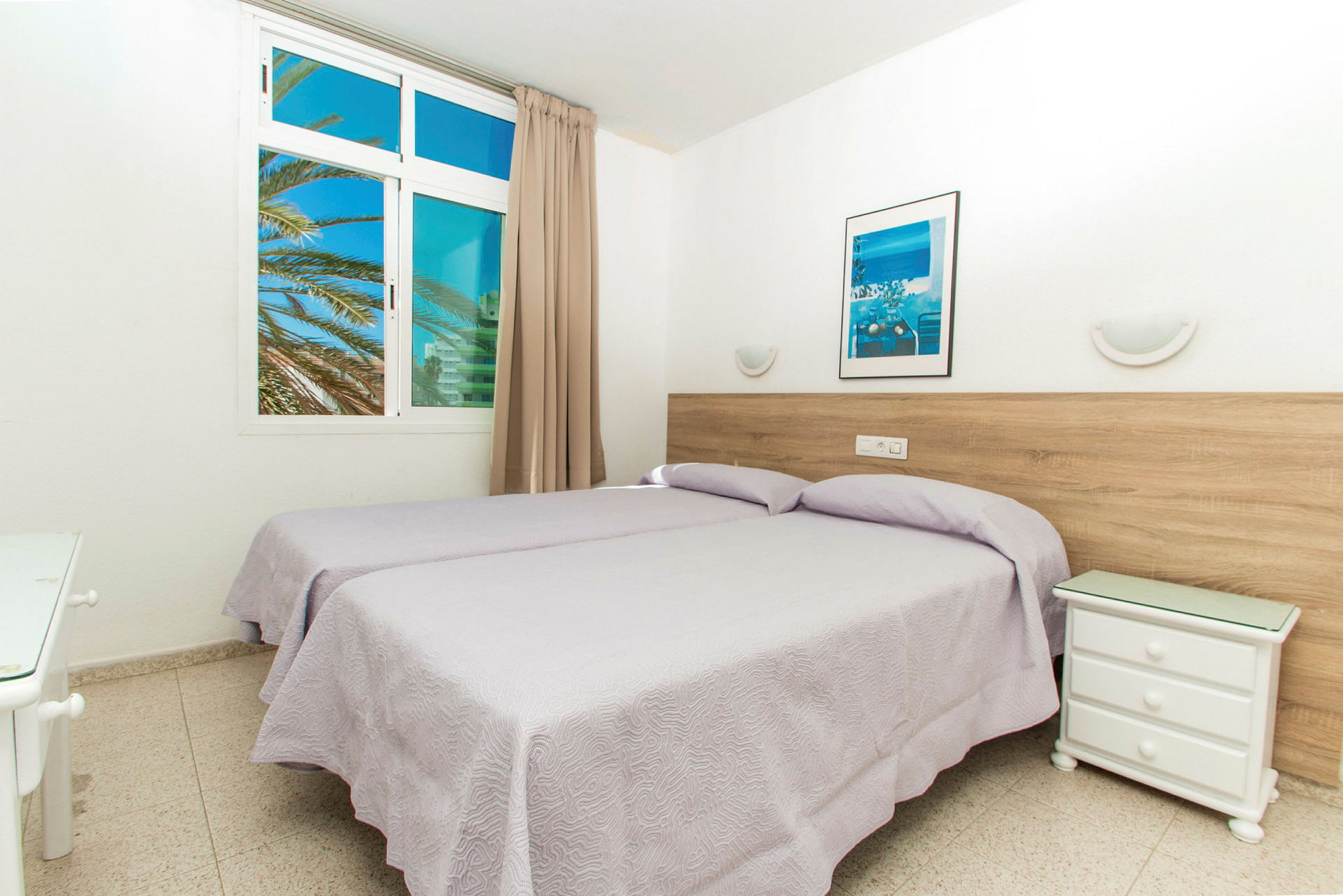 Ett hotellrum med två enkelsängar, ett nattduksbord och ett fönster med utsikt över palmer. (AI-genererad text)