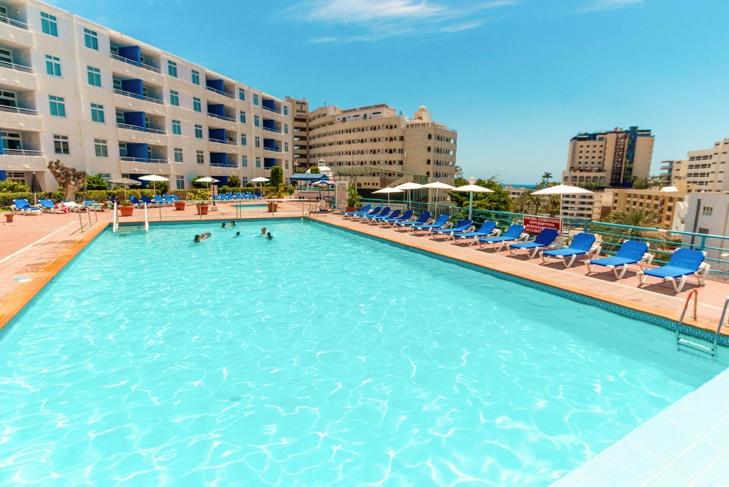 Stor swimmingpool utomhus omgiven av solstolar och hotellbyggnader. (AI-genererad text)