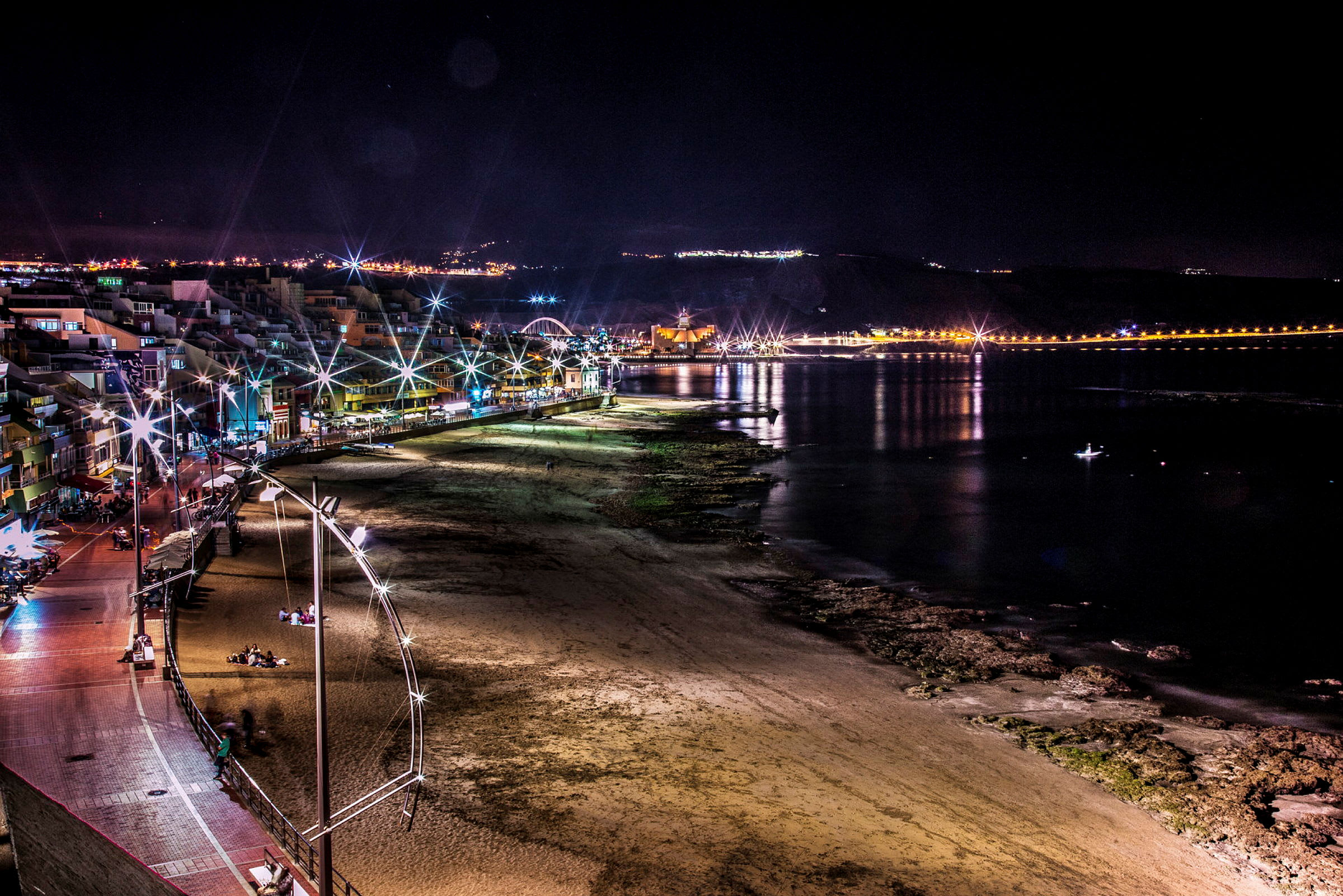 En strand och stad upplyst om natten, med reflektioner från ljusen i vattnet. (AI-genererad text)